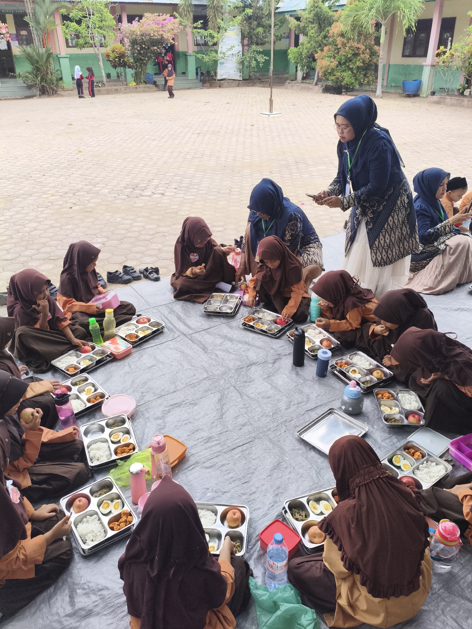 In the city of Baratpayaitek, Indonesia, a group of young girls in brown hijabs and orange uniforms are sitting in a circle on a gray tarp, eating lunch from metal trays. Some of the trays contain rice, boiled eggs, and vegetables. A few adults, also wearing hijabs, are present, observing the children. One adult, wearing a dark blue hijab and patterned dress, is holding a mobile phone. The scene appears to be an outdoor school setting, with a building and trees visible in the background. Shoes are neatly arranged on the ground, suggesting a communal gathering. The atmosphere is calm and orderly, with children focused on their meals.