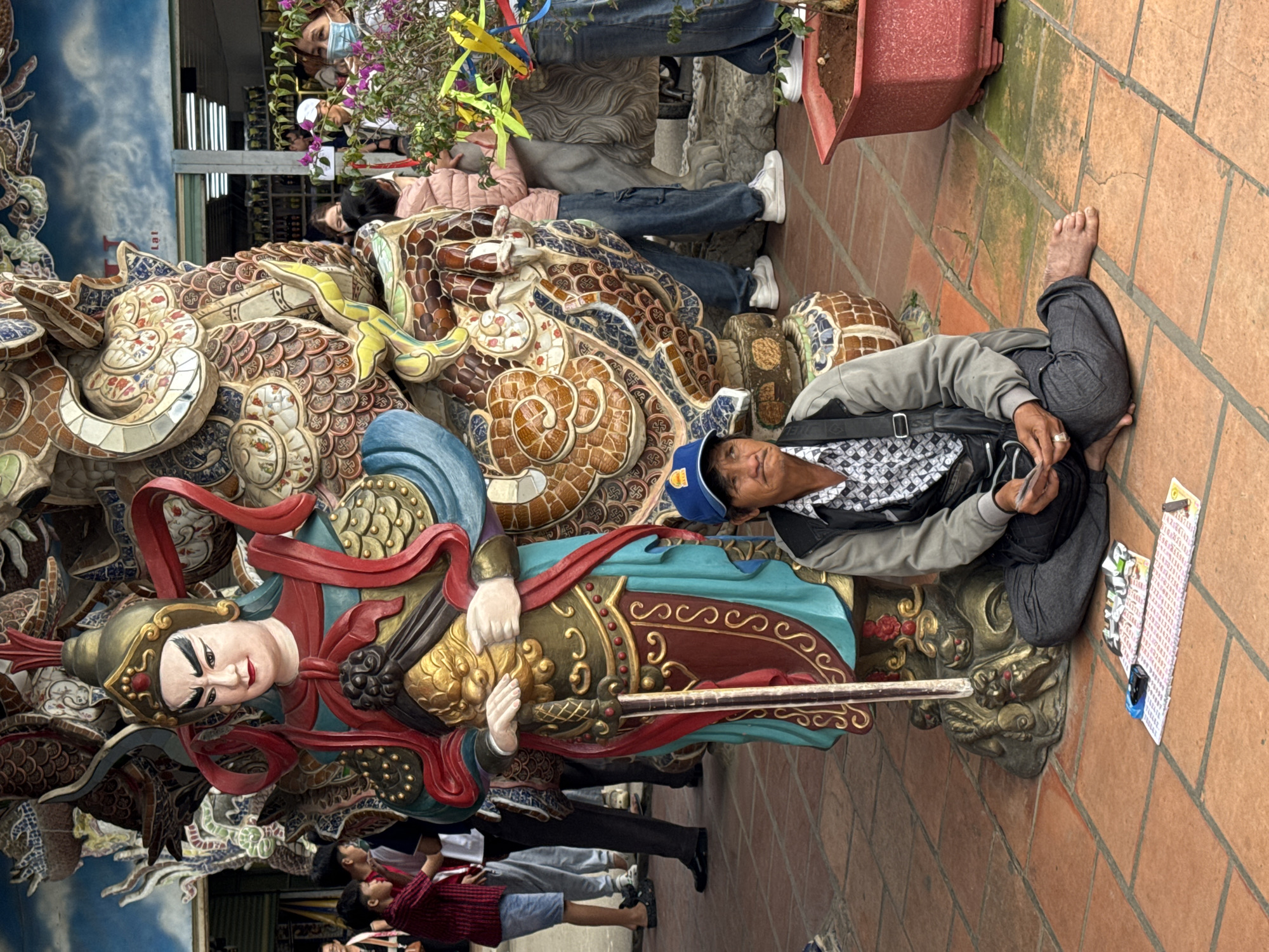 A man wearing a blue baseball cap, a checkered shirt, and a grey jacket sits on the ground in front of a large, colorful statue of a warrior in the city of Xuân Thành, Vietnam. The man is holding cards in his hands, and several decks of cards and lottery tickets are spread out on the ground in front of him. The statue is ornate, with intricate detailing and vibrant colors, depicting a warrior in armor holding a sword. Several other people, including children, are visible in the background, near other statues and structures, some wearing masks. The ground is paved with terracotta tiles. A red planter with a plant is visible to the right of the man.