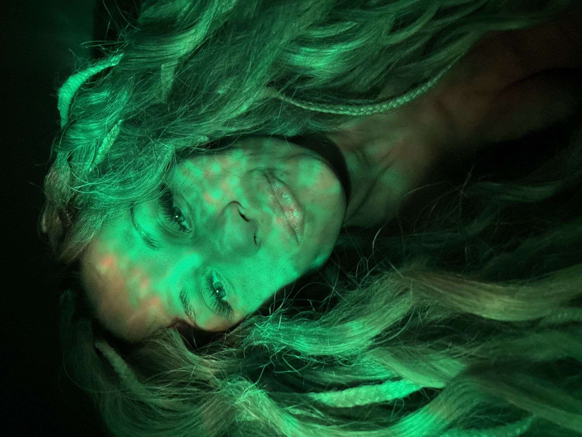 A close-up, low-angle shot of a person's face and hair, illuminated by green light. The person appears to be lying down, with their head tilted slightly. The hair is long, light-colored, and flowing around the face. There are distinct, braided strands incorporated into the hair. The person's eyes are closed. The skin has a mottled appearance due to the lighting, with areas of green and red hue. The background is dark, with no discernible objects or features. The image provides no indication of a location in Rusanovo, Russia or any other city. There is nothing to indicate any action or interaction.