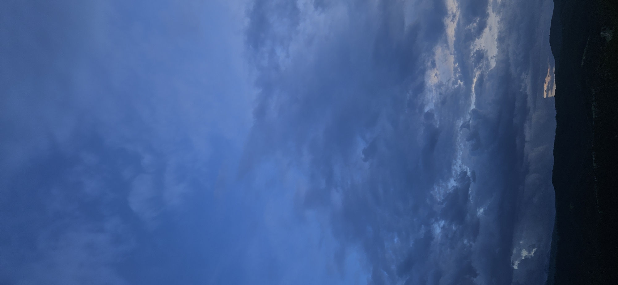 The image captures a dramatic sky and mountainous landscape, originally presented in a portrait orientation but depicting a horizon when viewed horizontally. The vast majority of the frame is dominated by an expansive sky, filled with a complex tapestry of clouds. These clouds exhibit a range of tones from deep, saturated blues and grays, hinting at dense atmospheric conditions, to areas illuminated with bright, almost white and pale yellow light. This contrast suggests either early morning dawn or late evening twilight, with the sun positioned low on the horizon, peeking through or reflecting off the cloud layers. Some cloud formations appear heavy and layered, creating a sense of depth and movement across the sky.
Below the dynamic sky, a dark, silhouetted landmass stretches across the bottom portion of the frame. This appears to be a range of mountains or heavily forested hills, their contours softened by distance and the fading light. A notable detail is a narrow, elongated band of intense, warm light that pierces through the clouds just above the mountain line, creating a striking luminous streak against the darker elements.
There are no people, objects, or visible activity within the scene, emphasizing the untouched natural beauty. The image evokes a sense of grandeur and tranquility, characteristic of mountainous regions. Given the context, this breathtaking natural vista is likely located in or around Gutsal, Bulgaria. No visible text is present.