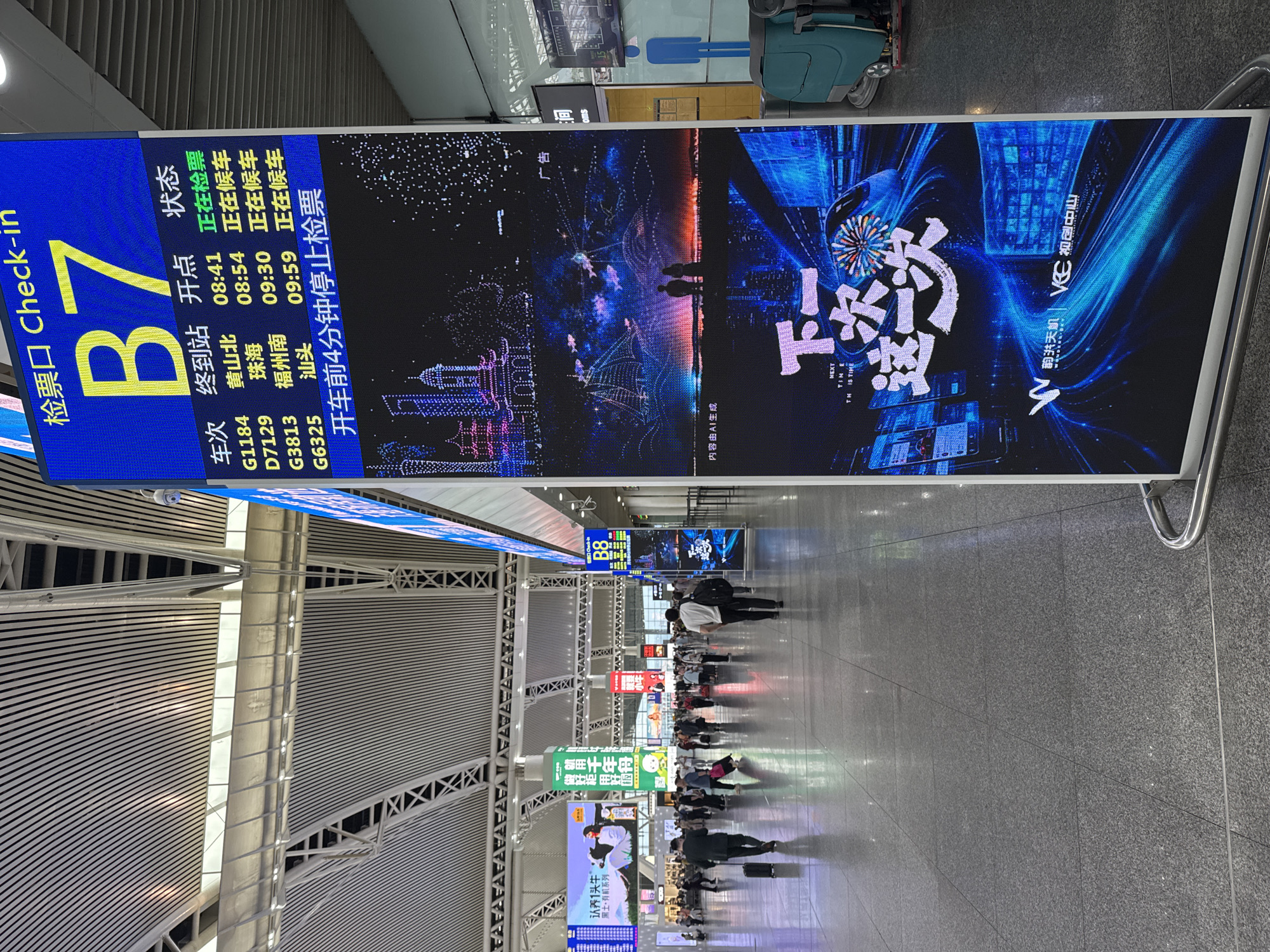 The image shows the interior of a train station in Panyu District, China. The scene features a long corridor with a high, arched ceiling made of metal beams. People are walking along the corridor, some with luggage. Overhead, digital signage displays information such as train departure times and check-in gates. One sign reads "B7" and lists departure information for various trains. Next to it, there's an advertising display, its images are generated by AI, featuring a promotional graphic for an advertisement. Other signs are visible in the distance, including one for "认养1头牛" (Adopt a Cow). The floor is shiny and appears to be made of gray granite. The lighting is bright and artificial.