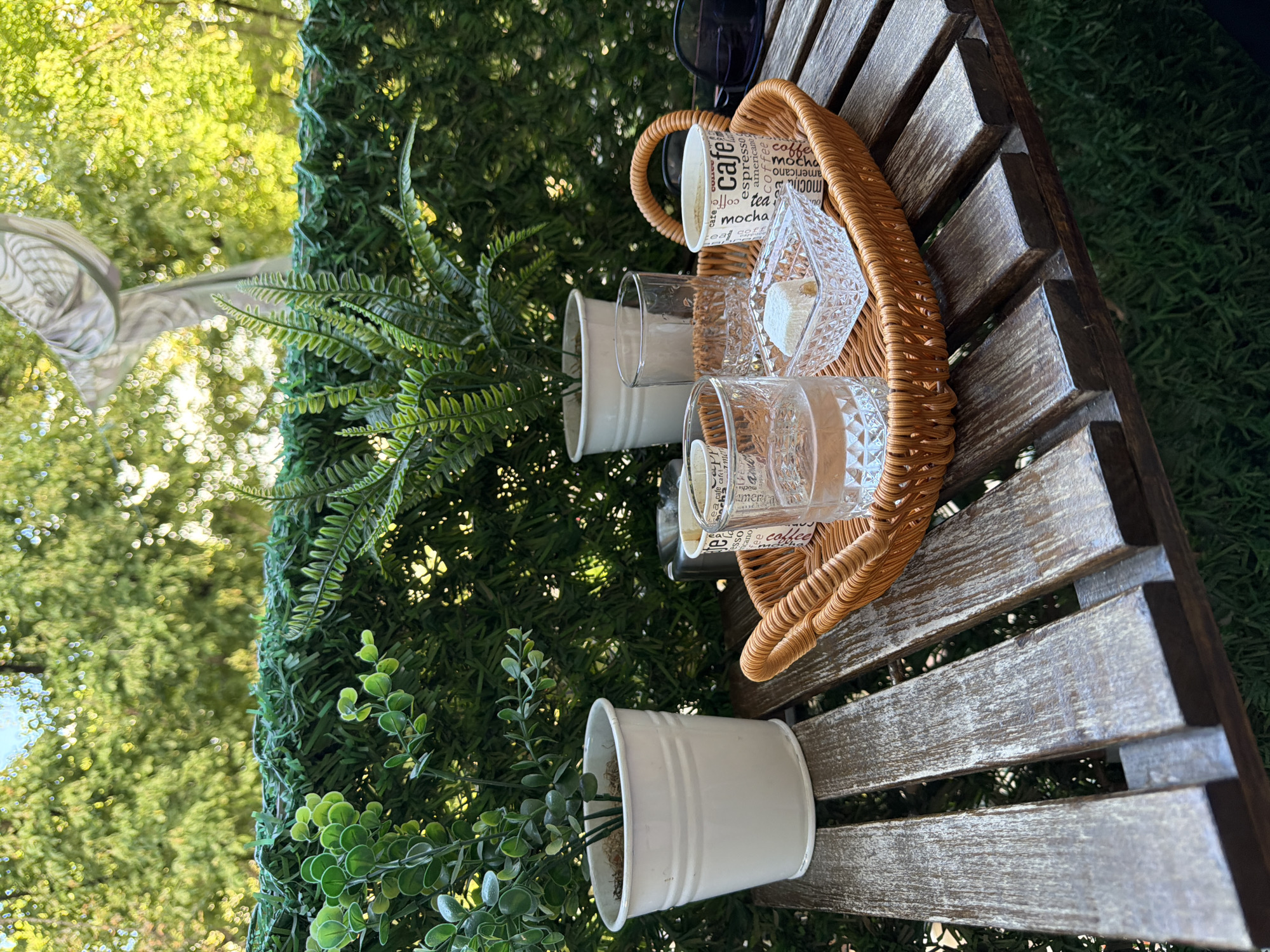 The image depicts a serene outdoor setting, likely a balcony or patio, in Kocaeli, Turkey. The focal point is a rustic wooden table laden with a woven basket. Inside the basket, there are two glasses of water, a small crystal dish containing a piece of Turkish delight, and a paper cup with the word "cafe" printed on it. Next to the basket, there is another empty glass and a white pot containing a lush green plant. In the background, a vibrant green hedge serves as a backdrop, with the blurred foliage of trees suggesting a sun-dappled environment. A hint of a statue or sculpture is visible through the greenery. The overall ambiance is one of quiet relaxation and enjoyment.