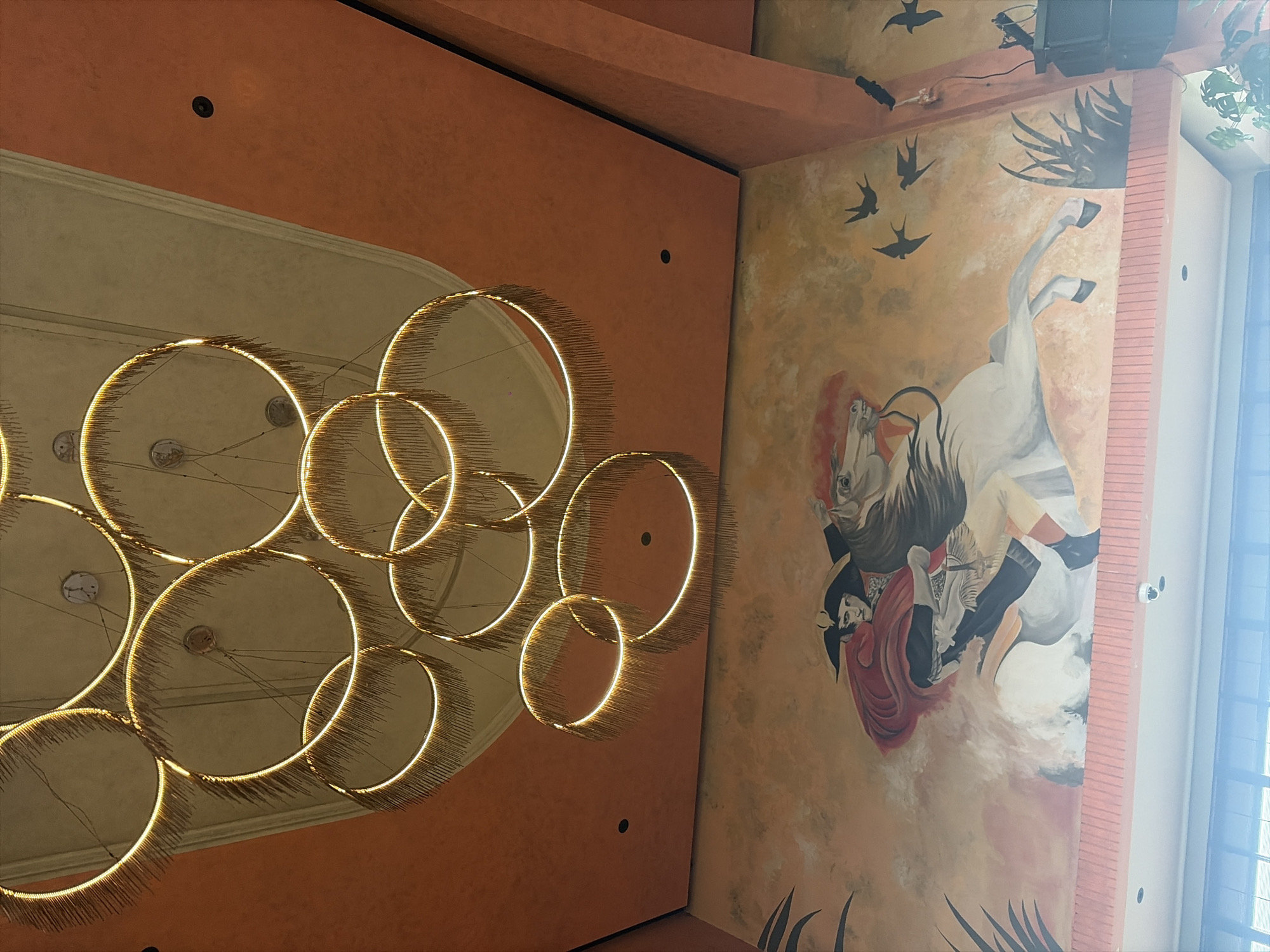 This image depicts a well-lit interior space with a prominent chandelier and a large mural. The chandelier is composed of multiple gold-colored rings with a fringe-like embellishment and is illuminated with warm light. The ceiling is painted in two distinct sections: a light beige area where the chandelier is mounted, and a vibrant orange section that extends to the right.
The mural, covering a significant portion of the wall, is a colorful depiction of a historical or mythological scene. It features a person in a red cape and a black hat, mounted on a white horse. The style of the mural suggests a classic artistic approach. Several black silhouettes of birds are scattered across the sky portion of the mural, adding to the dynamic composition. Palm-like foliage is also visible on the left and right sides of the mural.
The overall environment suggests a dining or entertainment venue, possibly a restaurant or hotel lobby, given the scale of the artwork and the decorative lighting. The lower part of the image reveals a hint of a window or glass partition with horizontal blinds, suggesting a connection to an outdoor or transitional space. A security camera is visible on a ledge below the mural. The presence of this artwork and the elaborate decor suggest a location that values aesthetics and possibly cultural themes. The location is identified as North Kuta, Indonesia.