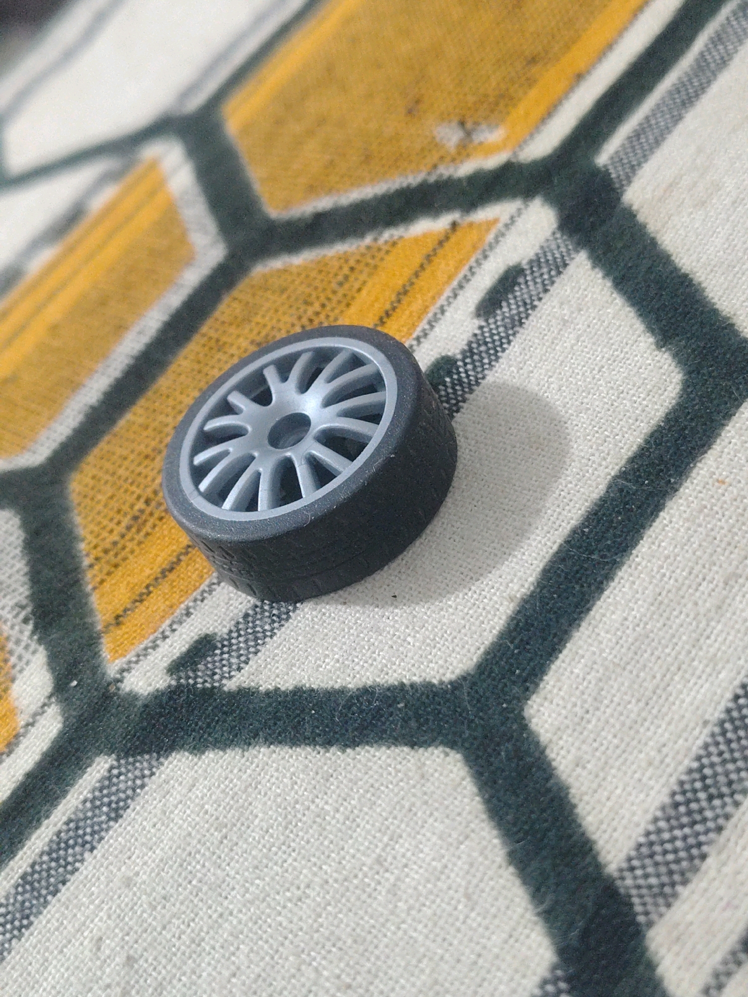 A single, miniature toy car wheel is positioned on a patterned fabric surface. The wheel has a dark grey tire and a silver alloy-style rim with multiple spokes. The fabric beneath is predominantly white with a pattern of interlocking hexagonal shapes outlined in dark grey, with some of these shapes filled with a yellow hue. The image is a close-up shot, with the wheel slightly off-center and casting a soft shadow. The texture of the fabric is visible, appearing to be a woven material, possibly a tablecloth or a blanket. There are no people or discernible environmental elements indicative of Trincomalee, Sri Lanka, present in the image.