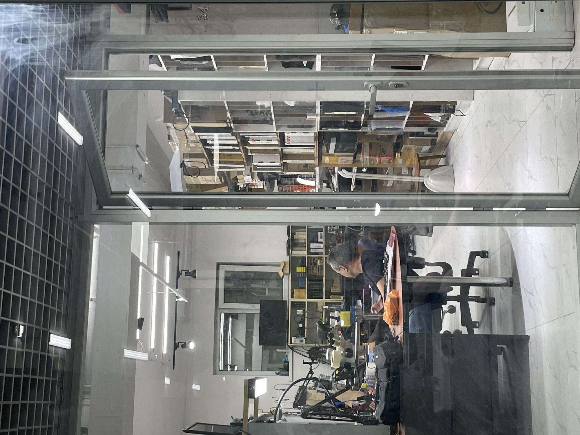 The image shows the interior of what appears to be an electronics repair shop or a similar business, viewed through a glass storefront. The scene is divided into two main areas by a doorway.
On the left, a man is seated at a cluttered workbench, facing away from the camera. He is wearing a dark shirt and blue jeans. The workbench is covered with various electronic components, tools, a microscope, a laptop, and a bag. The area behind him contains shelving units with more equipment and what looks like a window with blinds.
On the right, a brightly lit room is visible, filled with shelves stacked high with boxes, binders, and other items. The shelves extend from floor to ceiling. In the foreground of this area, a white dustpan and brush are leaning against a shelf. A door with a handle is slightly ajar.
The floor in both areas is tiled with a light-colored marble pattern. The ceiling features a grid of recessed lighting fixtures. The overall impression is one of a working environment with a significant amount of stored inventory. The presence of what appear to be shipping boxes and various electronic parts suggests a business involved in sales or repair of such items. There are no overt clues within the image that definitively pinpoint the location to Almaty, Kazakhstan, but the scene is consistent with a retail or service business.
