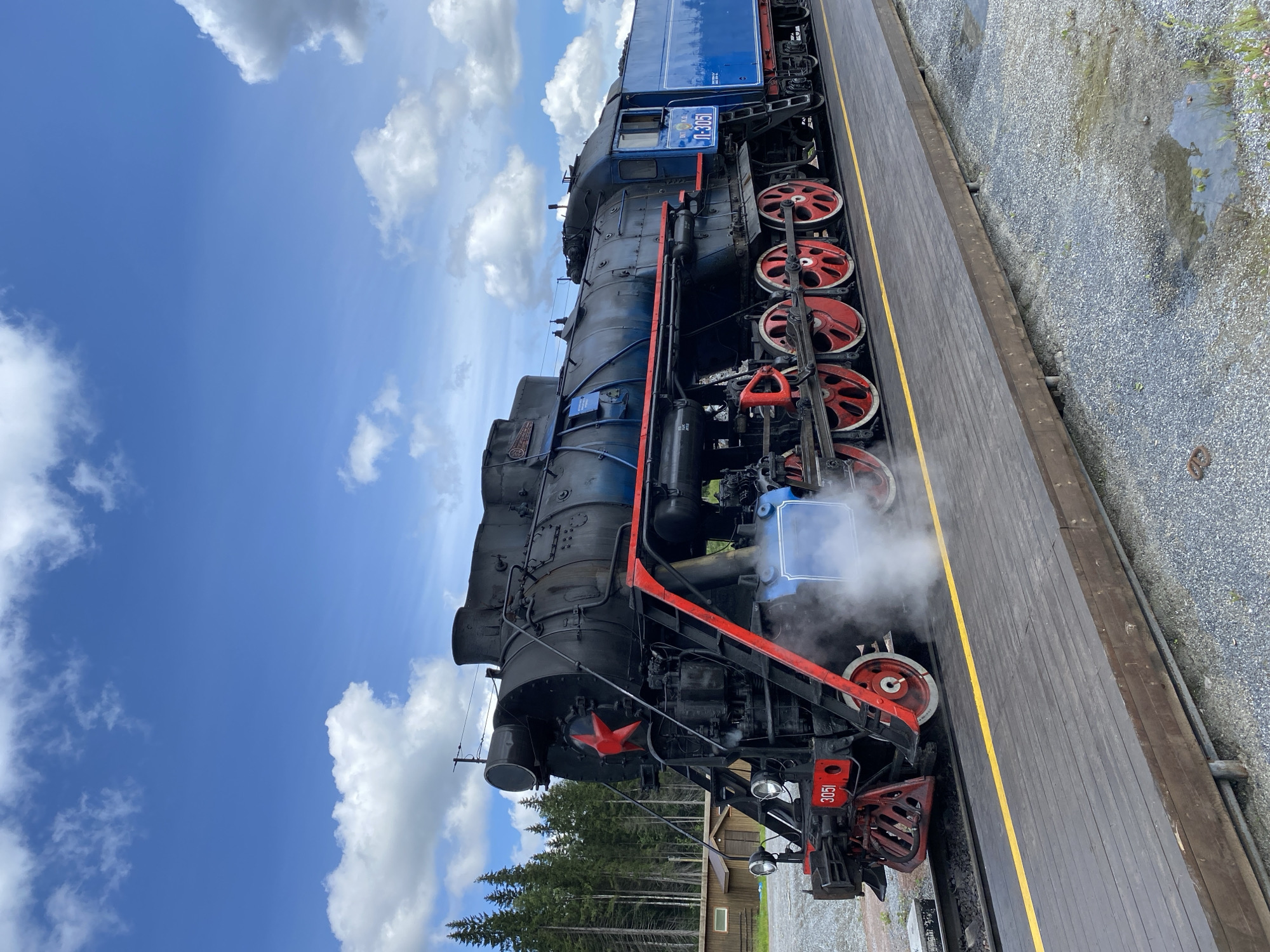 A vintage black and blue steam locomotive, numbered "3051", is parked on a wooden platform with a yellow stripe bordering a railway track in Ruskeala, Russia. The locomotive has a prominent red star on its front and red-rimmed wheels. Steam is emanating from the engine. In the background, a dense forest of evergreen trees is visible, along with a wooden building. The sky is a bright blue with scattered white clouds. The ground around the track is covered in gravel. The image is a medium shot, capturing the side profile of the train.