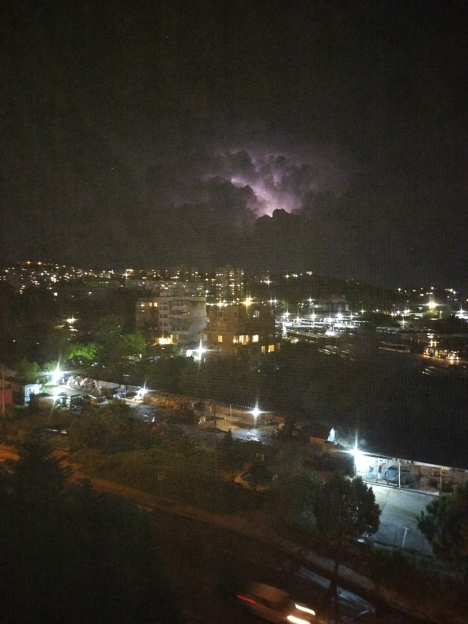 The image depicts a nighttime cityscape with a lightning storm in the background, likely taken in Varna, Bulgaria. In the foreground, there are trees and roads. A vehicle with blurred headlights is visible in motion along the road. Middle ground reveals buildings and a bus station, illuminated by streetlights and lights from the buildings. The background shows a city skyline with numerous lights, topped by a dramatic lightning storm in the clouds.