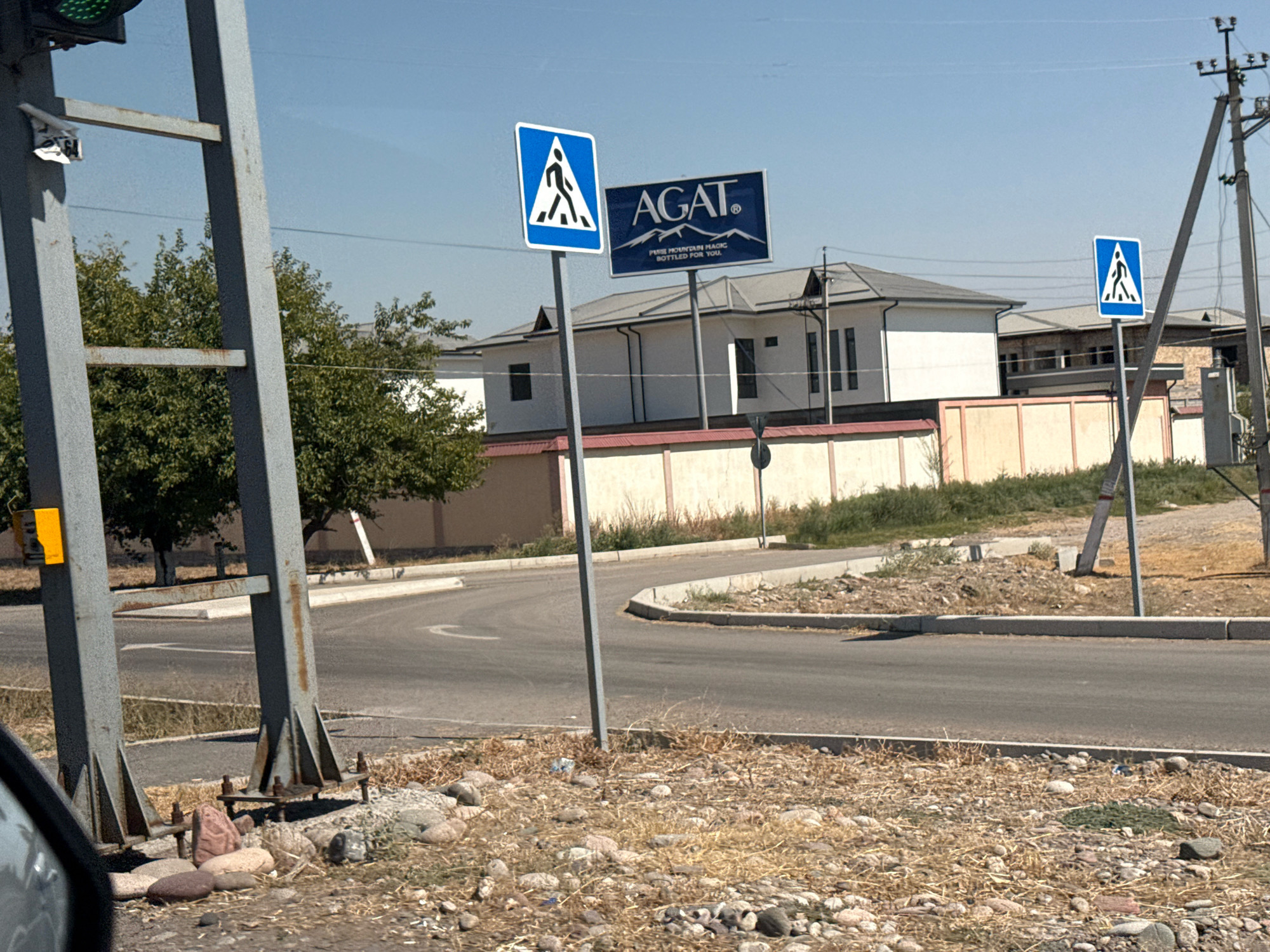 The image captures a roadside scene in Tashtugizak, Uzbekistan, on a clear, sunny day. A traffic light, partially visible at the top left, is mounted on a gray metal pole. Adjacent to it, a larger gray metal structure with horizontal supports is present. A blue and white pedestrian crossing sign, depicting a silhouette of a person walking on a crosswalk, stands on a gray pole. To its right, a dark blue rectangular billboard displays the brand "AGAT" in white lettering, with an image of mountains and the tagline "PURE MOUNTAIN MAGIC BOTTLED FOR YOU." Further to the right, a utility pole with crossbars and wires is visible.
The environment consists of a paved road that curves to the left, bordered by a concrete curb. Dry grass, pebbles, and rocks are scattered on the roadside. In the background, a beige fence runs along the property line in front of a white building with a dark roof. Trees with green foliage are also present in the background. The scene appears to be an intersection or a road leading into a residential or commercial area. There are no people or vehicles actively engaged in actions within the frame.