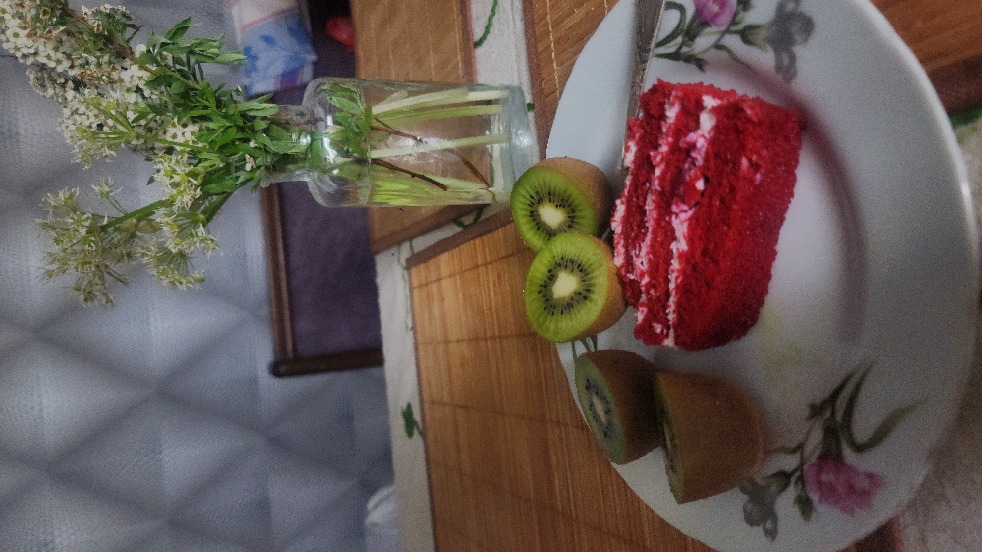 A top-down, slightly tilted view shows a still life on a table covered with a light-colored cloth and a bamboo mat. On the left, a clear glass vase holds white flowers and green foliage. To the right, a white plate with floral patterns contains a slice of red velvet cake with white frosting and three kiwi fruits, one cut in half. The scene's exact location within Moscow, Russia, cannot be determined from the provided image.