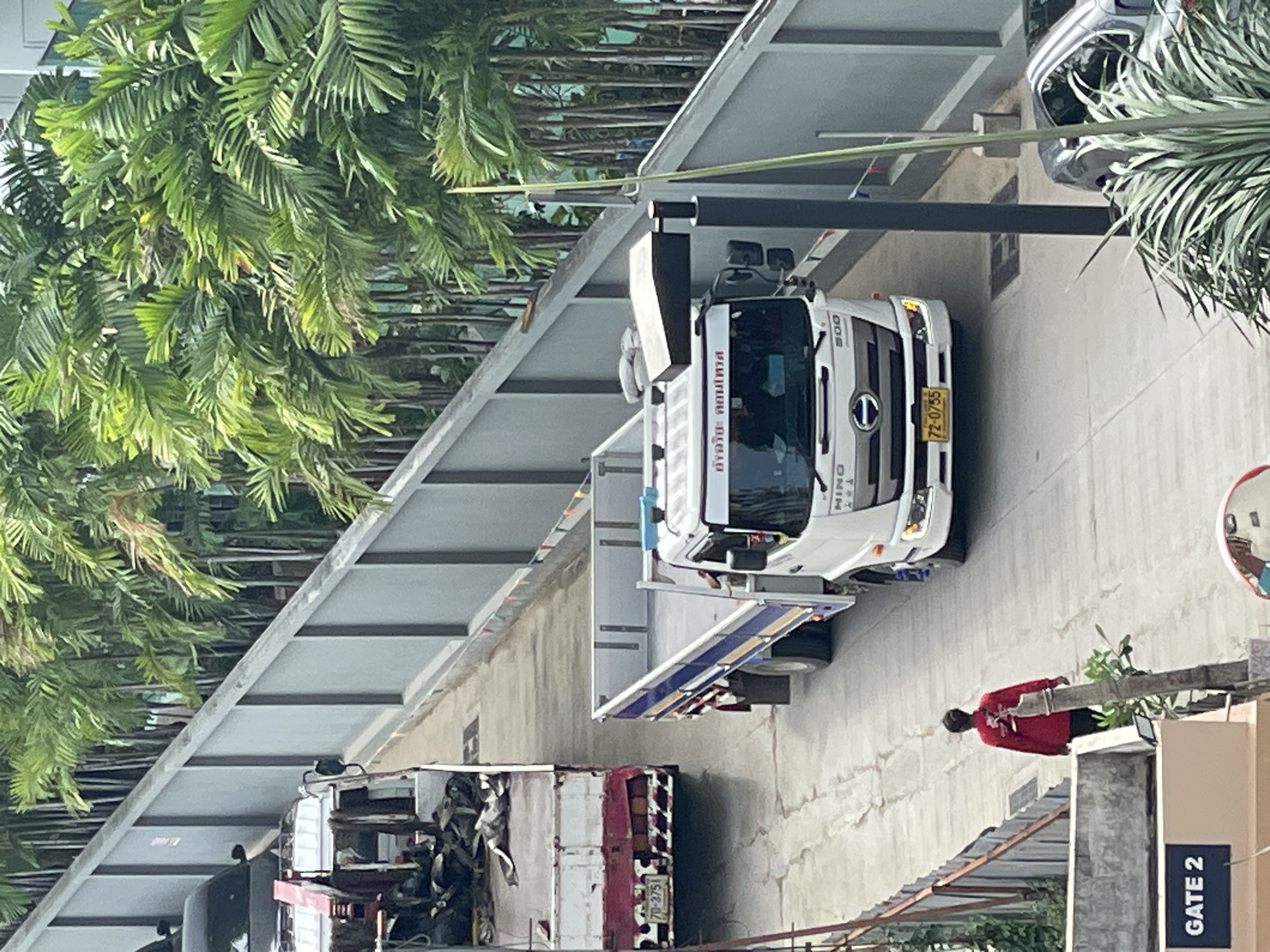 A white Hino truck with a blue and yellow stripe on its side is driving up a ramp. The truck has a license plate that reads "72-0755". To the left of the Hino truck is another truck with a red and white cab, which is also driving up the ramp. The truck has a license plate that reads "70-3751". A person in a red shirt is walking on the ramp. In the background, there are palm trees. The scene appears to be in an urban environment, possibly a city like Pattaya City, Thailand, indicated by the presence of palm trees and signage.