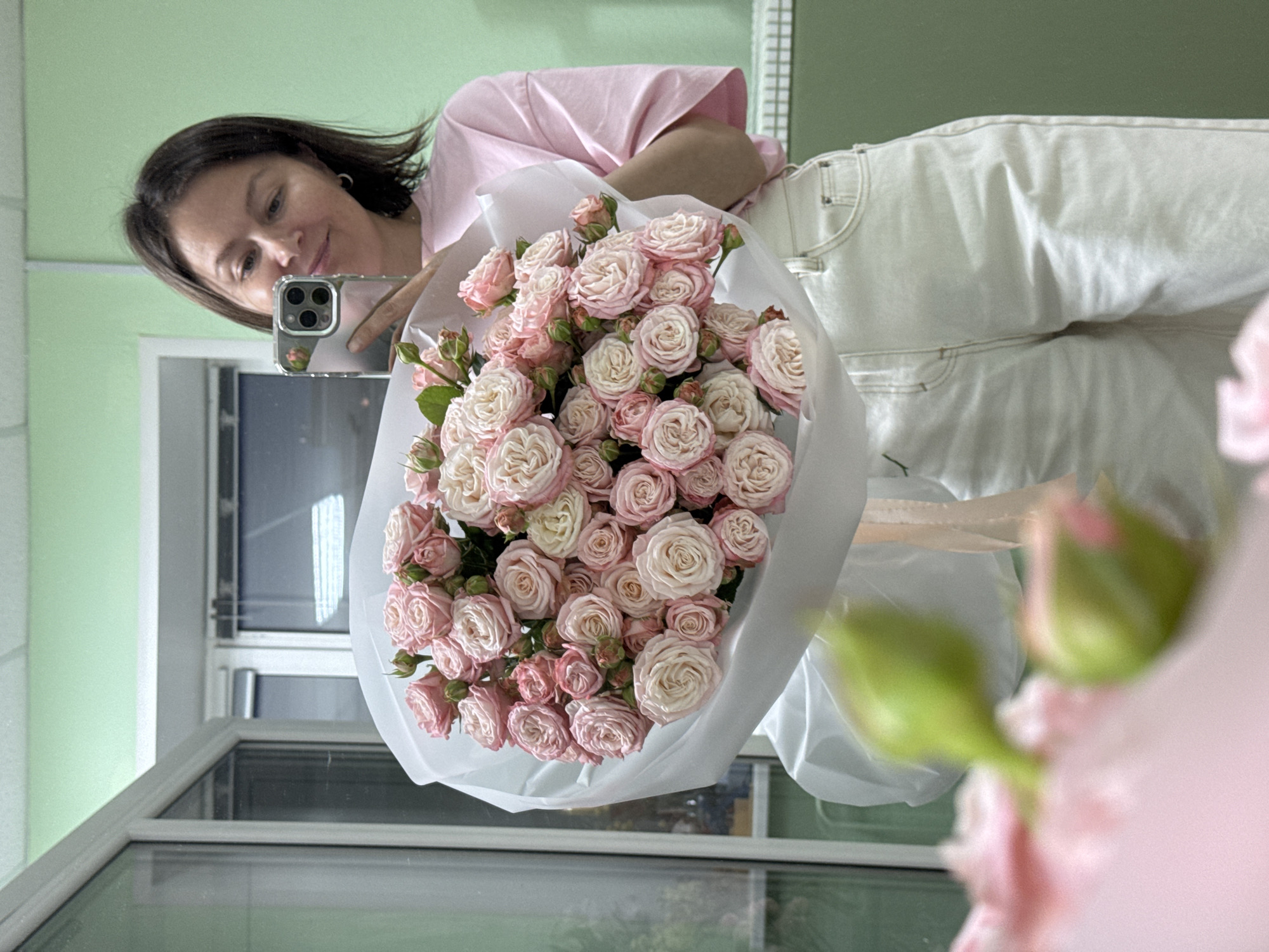 A young woman with brown hair, wearing a pink t-shirt and white jeans, is taking a selfie in a mirror. She is holding a large bouquet of light pink and white roses. The background appears to be an indoor space with light green walls and a window or doorway on the left. The image is a mirror reflection, indicated by the woman holding her phone to capture the shot. The location is not determinable from the image.