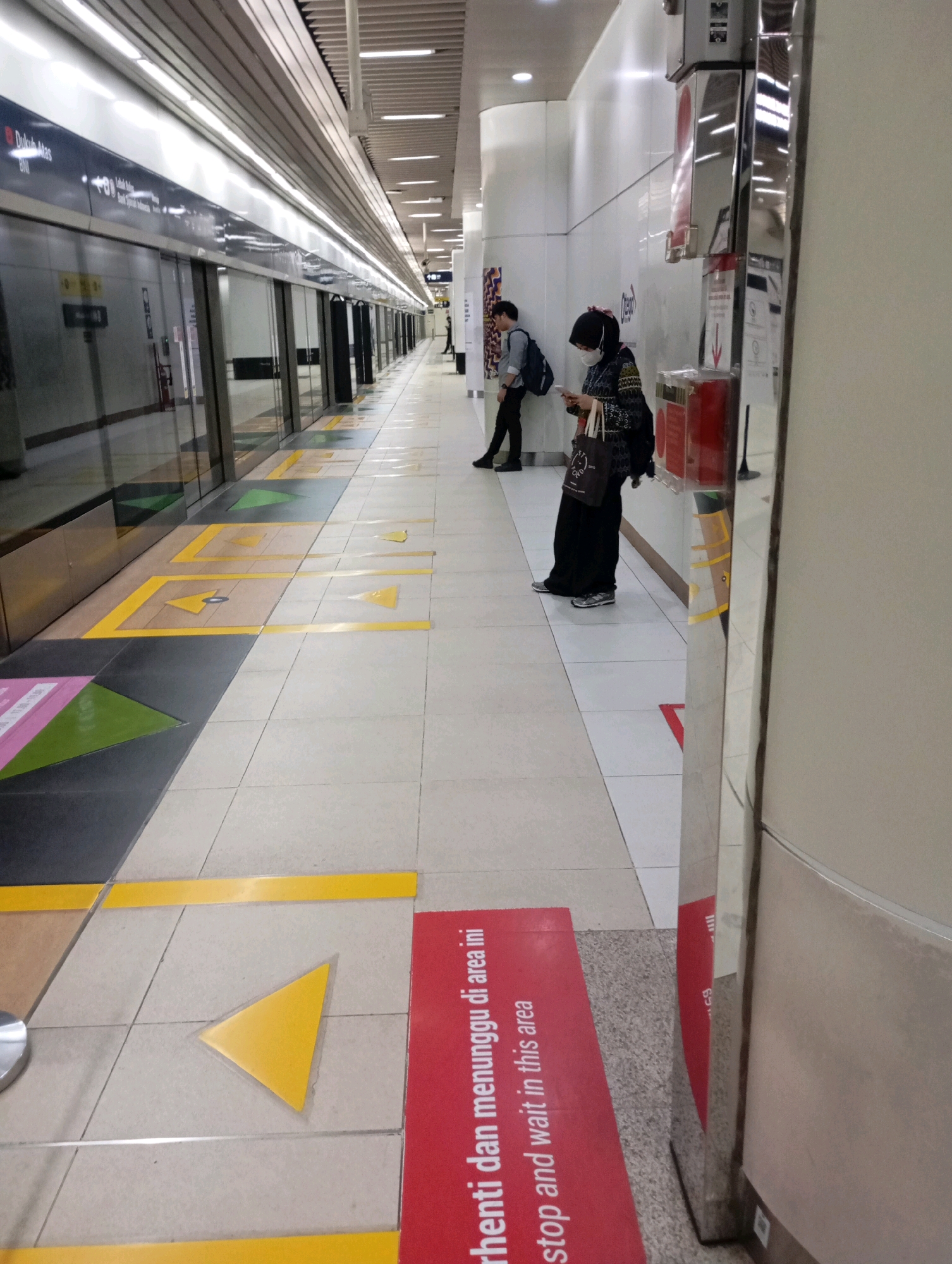 The image shows a subway station platform in Central Jakarta, Indonesia. The platform is clean and modern, with tiled floors and automatic doors lining the track. Yellow directional arrows are painted on the floor, guiding passengers. Two individuals are visible: a man in a light blue shirt and black pants leaning against a wall, and a woman in a headscarf and patterned top, wearing a mask and looking at her phone while holding a bag. The woman is standing on a red mat with text in both Indonesian and English that reads "Berhenti dan menunggu di area ini" and "Stop and wait in this area." The scene is well-lit by overhead fluorescent lights. The overall impression is one of an orderly and functional public transportation space.