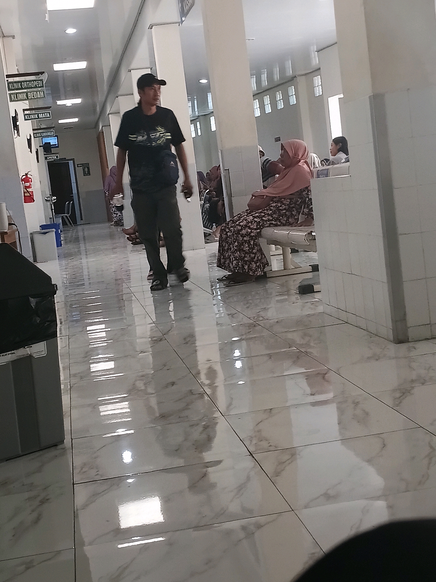 The image depicts a hallway in a public building, possibly a hospital or clinic, with polished marble floors reflecting overhead fluorescent lighting. On the left wall, there are several signs indicating different departments, including "KLINIK ORTHOPEDI," "KLINIK BEDAH," and "KLINIK MATA." A man wearing a black t-shirt, dark pants, and a black baseball cap is walking down the hallway, looking to his right. He is carrying a dark object in his right hand. Further down the hallway, on the right side, a woman wearing a light pink hijab and a patterned dress is sitting on a bench, facing away from the camera. Another woman, partially visible, is seated next to her. The environment appears to be clean and well-maintained, with the polished floor indicating recent cleaning. The overall impression is of a busy waiting area or corridor within a healthcare facility. The presence of specific clinic names suggests this location is in Indonesia, likely within a city like Purwodadi, given the context of the prompt.