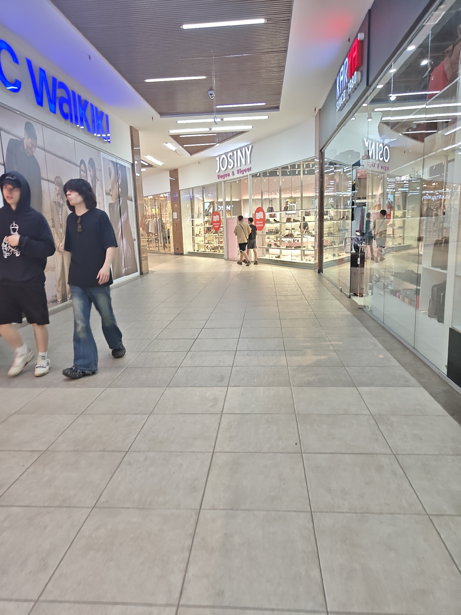 Two young men are walking on a tiled floor inside a modern shopping mall. The man on the left is wearing a black hoodie, black shorts, and white sneakers. He is facing forward with his head slightly down. The man on the right is wearing a black t-shirt, blue flared jeans, and dark shoes. He is also facing forward, looking slightly to his right. Behind them, on the left side of the image, a large blue sign with white lettering reads "WALKIKI". Further down the mall, a store with a sign reading "JOSINY" displays shoes and handbags. On the right side of the image, another store has a red sign that says "KARYA SINCE 1980". The mall environment features a suspended ceiling with integrated lighting and large glass storefronts showcasing merchandise. The floor is made of large, light-colored square tiles. Based on the "WALKIKI" store, this scene is likely located in the Family Park shopping mall in Petropavl, Kazakhstan, as this is a known retailer there.