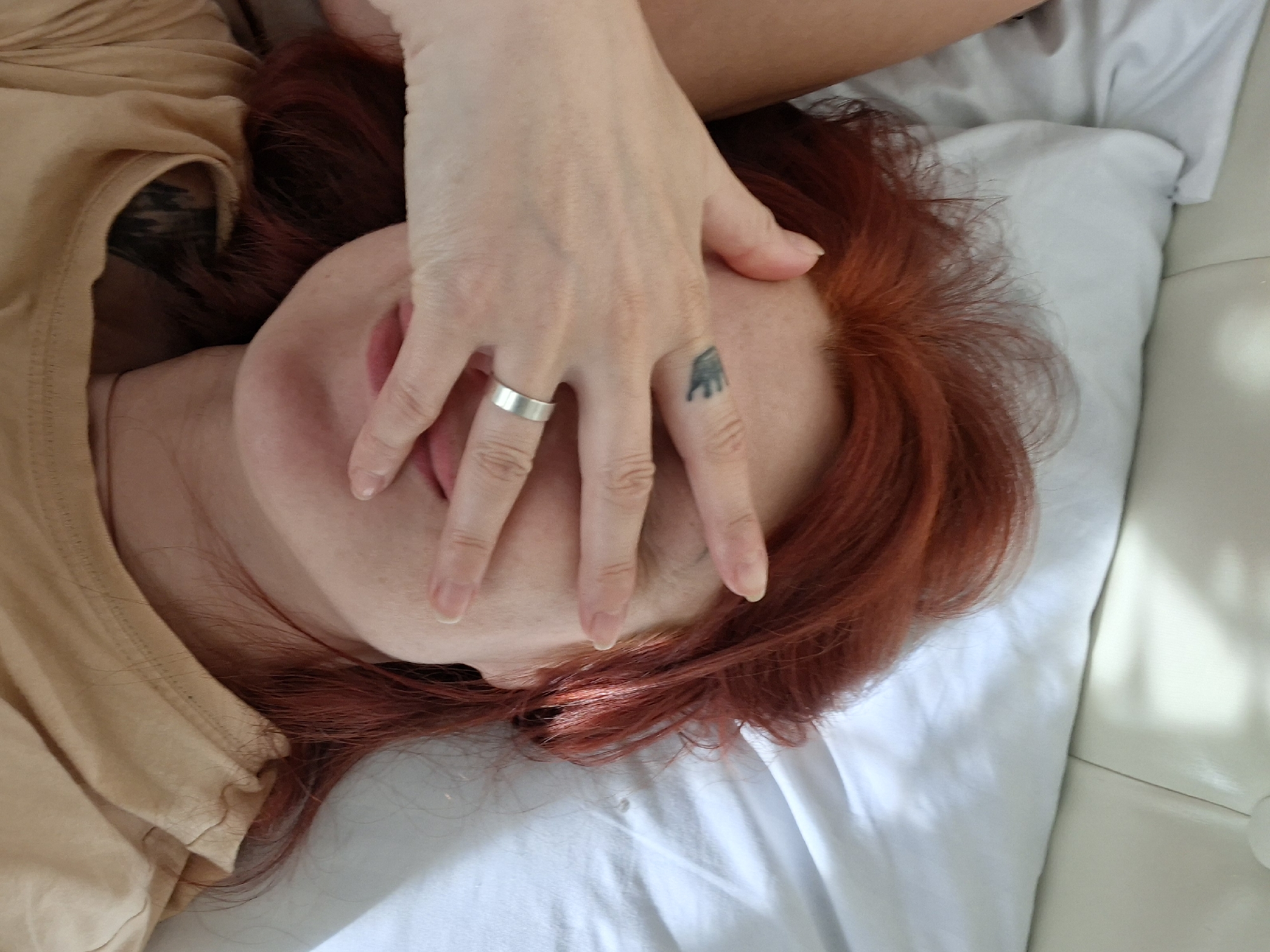 An overhead shot shows a person lying on a white bedspread. The person has vibrant red hair fanned out around their head. Their right hand is raised, obscuring their eyes and the upper part of their face. A silver ring is visible on the ring finger of the hand, and a small tattoo is visible on the knuckle of the middle finger. The person is wearing a light tan t-shirt, and a tattoo is visible on their upper chest. The background consists of the white bedspread and a white headboard. The image appears to be taken indoors. There is no indication of the location being in the city of Myadzyel, Belarus.