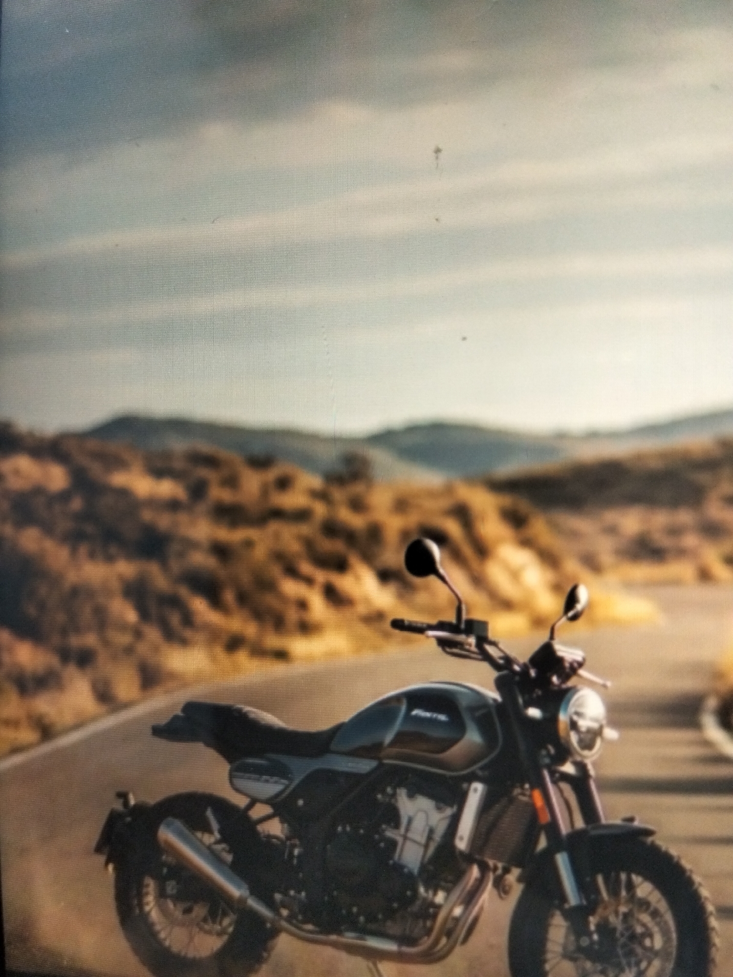 A dark gray motorcycle is parked on a paved road that curves through a barren, hilly landscape. The sky above is overcast with pale gray clouds. The motorcycle is the primary subject, positioned in the foreground and taking up a significant portion of the frame. It is facing towards the right of the image. The road appears to be a rural route, with dry, brown vegetation and hills stretching into the background. The lighting suggests it might be late afternoon or early morning, with a soft, diffused light illuminating the scene. There are no people visible in the image, and no actions are taking place beyond the static presence of the motorcycle. The environment is arid and undeveloped, characteristic of a remote or undeveloped area. There is no indication of the city of Payagaboh, Indonesia.