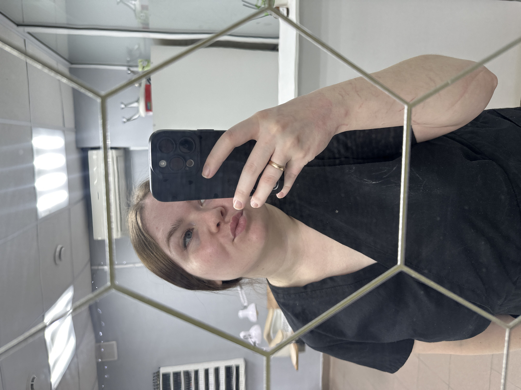 A person is taking a selfie in a mirror with a hexagonal frame. The person is wearing a black shirt. The reflection in the mirror shows a room with white tiles and a radiator. The city of Chaikovsky, Russia, is not discernible from the image.