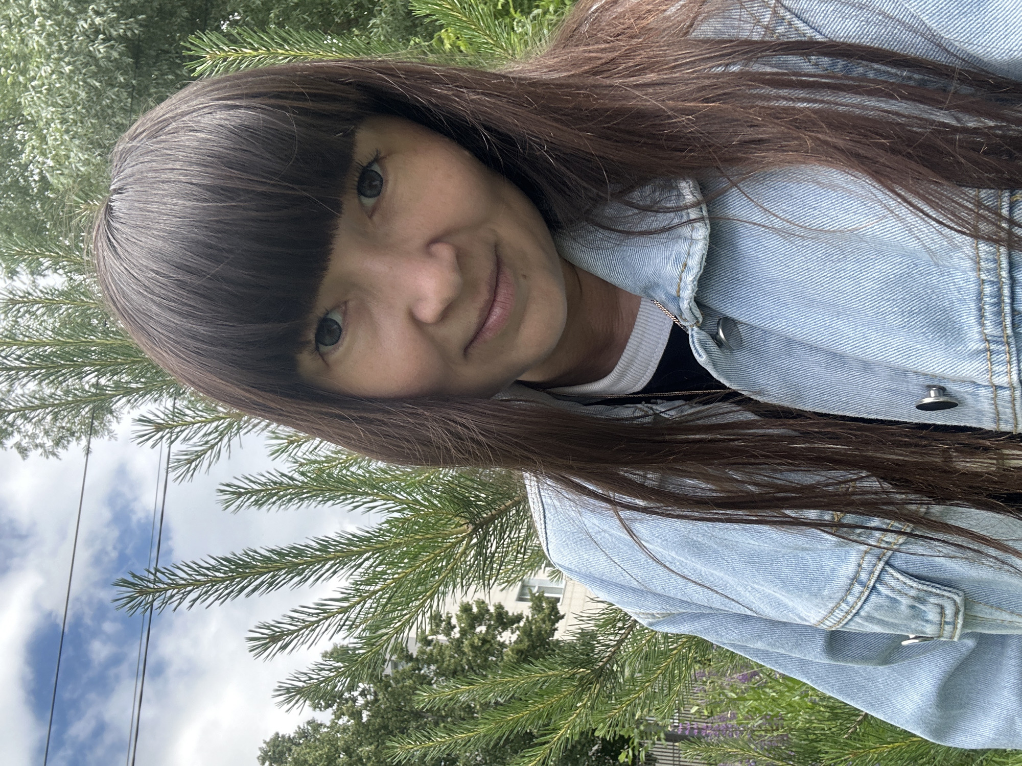 A medium shot selfie features a woman with long, dark brown hair and bangs, wearing a light blue denim jacket over a black and white top. She has a light complexion, dark eyes, and is looking towards the camera with a slight smile. The background consists of lush green pine branches and trees, with a partially visible building and cloudy sky in the distance. The setting appears to be outdoors, possibly in a park or residential area in Chaikovsky, Russia, given the presence of trees and the architectural style of the building.