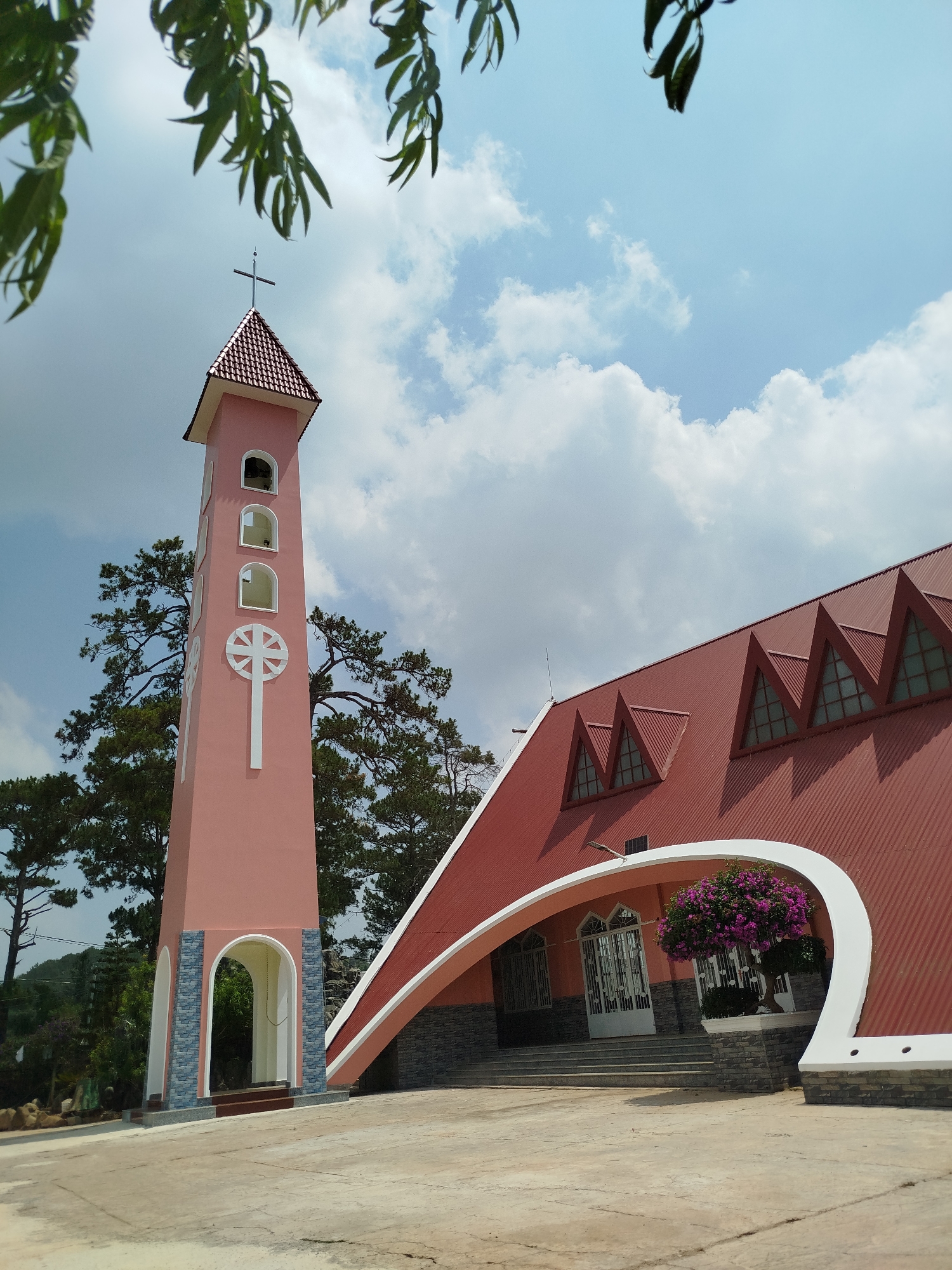 The image depicts the Domaine de Marie church in Đa Thọ, Vietnam. The church features a pink bell tower topped with a cross and a red tiled roof with distinctive triangular dormer windows. An arched white structure supports the roof above the entrance. A tree with purple flowers sits in front of the entrance. The scene is captured during the day under a partly cloudy sky. Green tree branches are visible in the upper left corner.