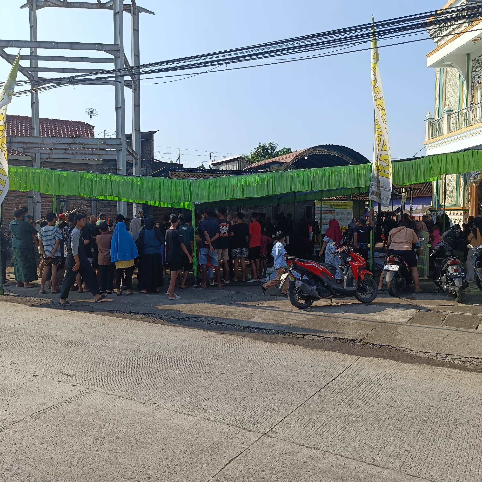 A large group of people are standing under a green awning in Sukomulyo, Indonesia. A building under construction with a metal frame is visible in the background. Several motorcycles are parked nearby. The street in front of the crowd is paved with concrete.
