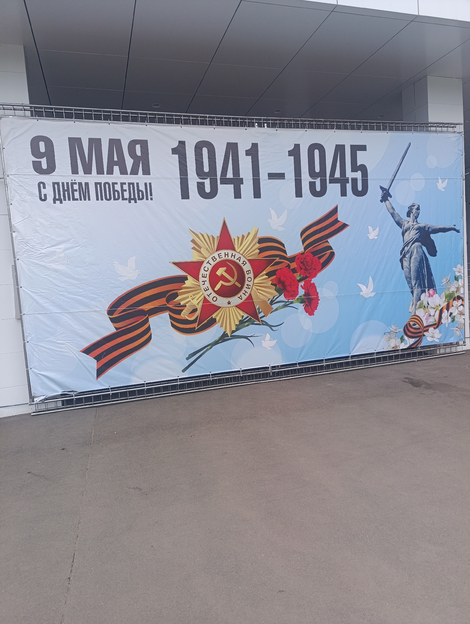 A banner hangs on a metal railing in the city of Unknown, Unknown. The banner displays the text "9 МАЯ С ДНЁМ ПОБЕДЫ!!" and "1941-1945." It features images of a Soviet star medal, a ribbon, carnations, a statue of a woman with a sword, and white doves. The background of the banner is light blue. The banner is attached to a building with white siding. The ground in front of the banner is asphalt.