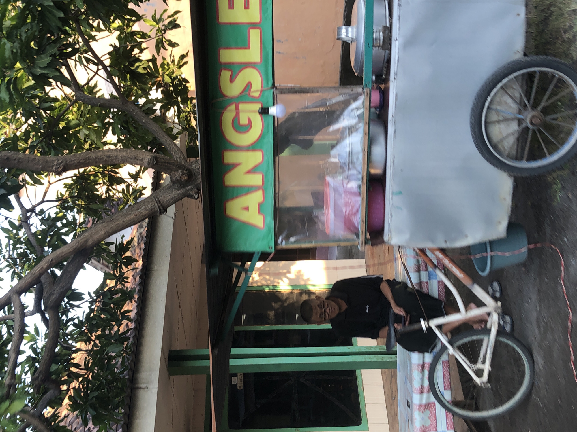 A man wearing glasses and a black polo shirt sits on a bench next to a food cart in Kalidawir, Indonesia. The food cart has a green banner with yellow lettering that reads "ANGSLE". A bicycle is parked in front of the cart. The man is holding a smartphone. A blue bucket is attached to the side of the food cart. In the background, there is a building with a tiled roof and a tree with green leaves. The sun is shining, casting shadows on the ground.