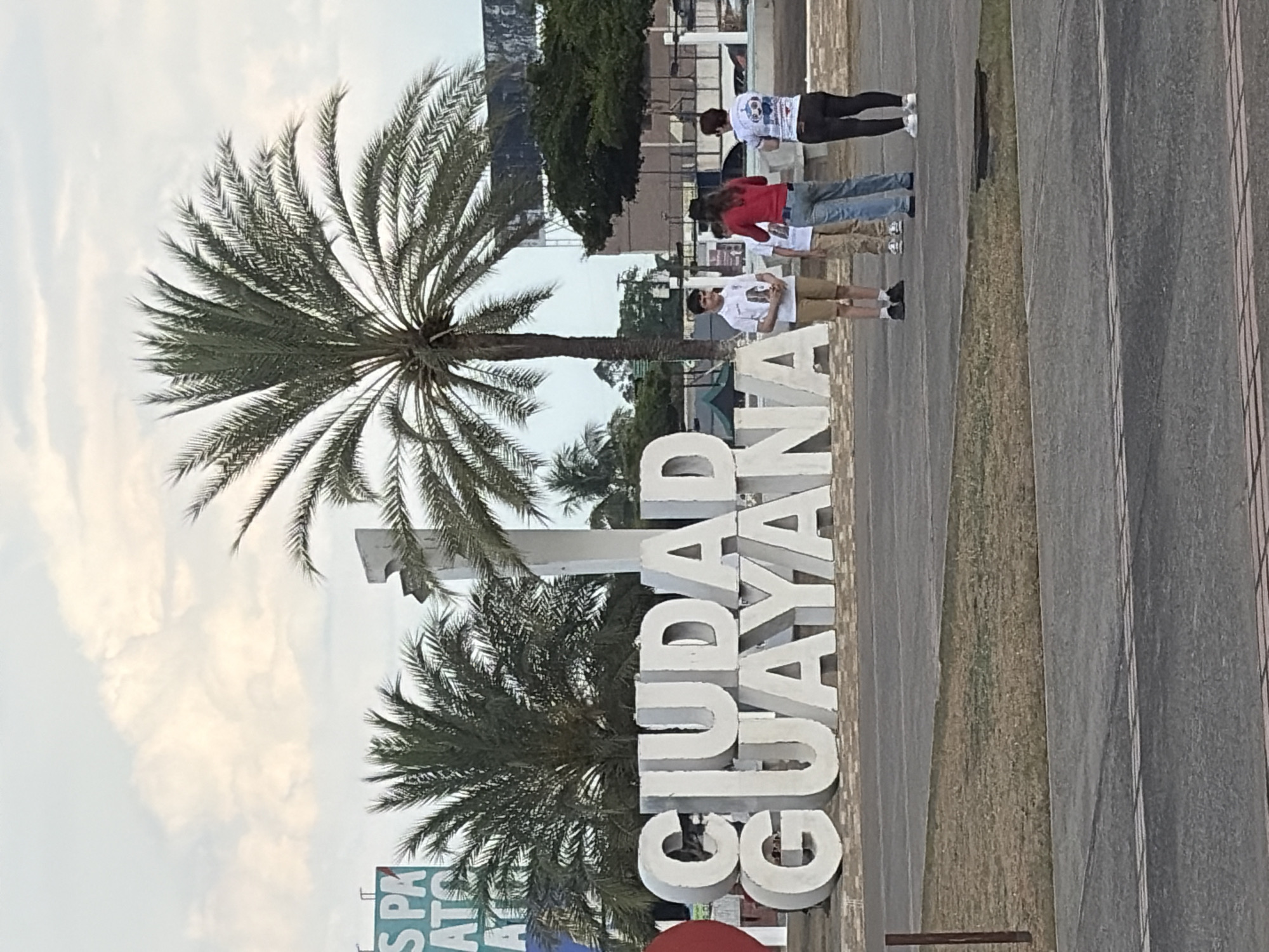The image shows the "Ciudad Guayana" sign in Guayana City, Venezuela. The large white letters are situated on a paved area, likely a public space. A palm tree is visible behind the sign.
Four individuals are present, positioned near the sign. From left to right:
- A person in a white t-shirt and khaki shorts, wearing sunglasses.
- A person in a red shirt and blue jeans.
- A person in a white t-shirt and blue jeans.
- A person in a white t-shirt with black leggings or pants.
The area around the sign is a mix of paved surfaces and vegetation. A building is seen in the background. The sky appears cloudy.