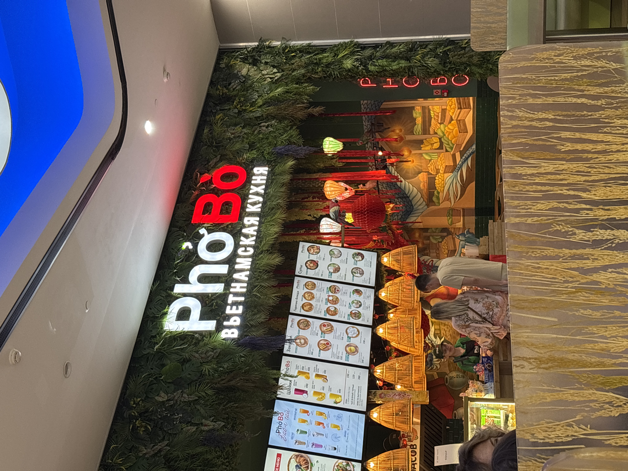 The image shows the exterior of a Vietnamese cuisine restaurant named "PhoBo" in Takhtamukai, Russia. The restaurant's facade is decorated with lush green artificial foliage. The prominent "PhoBo" logo is illuminated in white and red neon lights, with the text "ВЬЕТНАМСКАЯ КУХНЯ" (Vietnamese Cuisine) underneath. Several illuminated menu boards are displayed, showcasing different dishes and prices in Russian. Hanging from the ceiling are decorative bamboo lamps and traditional Asian lanterns, including red paper lanterns and green lotus-shaped lanterns. A mural depicting a dragon and stalls filled with produce is visible on the right side of the facade.
In the foreground, a counter is staffed by two employees wearing green aprons and caps with the "PhoBo" logo. A female customer in a floral print shirt is interacting with the staff at the counter, while a male customer stands nearby. Several other customers are partially visible in the foreground, either seated or waiting. The interior seating area features tables covered with a textured material resembling dried wheat stalks. The overall environment suggests a bustling food establishment within a modern shopping mall, indicated by the ceiling structure and lighting.