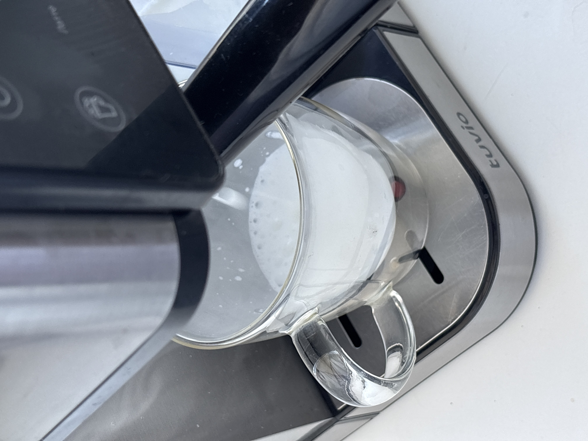 A close-up shot shows a clear glass mug filled with frothed milk sitting on the drip tray of a stainless steel and black espresso machine. The milk has a light foam on top. The espresso machine has some buttons with icons on the right side, including what appears to be a coffee cup. The brand name "tuvio" is visible on the drip tray. The background is a plain white surface. The image does not provide any information about the city of Perm, Russia.