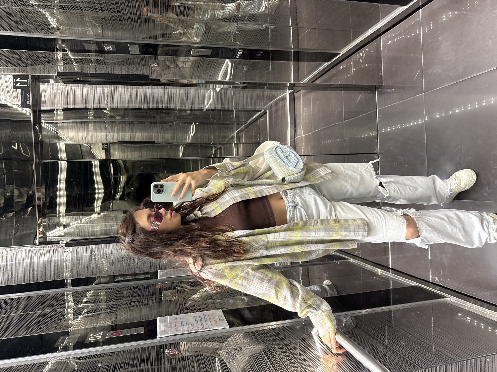 A young woman stands in a mirrored elevator, capturing a selfie with her phone. She is dressed in ripped, light-wash jeans, a brown top, and a plaid yellow and white jacket. Her brown, wavy hair is held back with a clip. She also wears sunglasses. The elevator's interior features shiny, dark tiled floors and mirrored walls, creating reflections and depth. Lines of decorative strings hang vertically, adding texture to the space. A small, light-blue bag is slung across her body. Elevator floor numbers are visible, with signage on the walls. The location is in Unknown, Unknown.