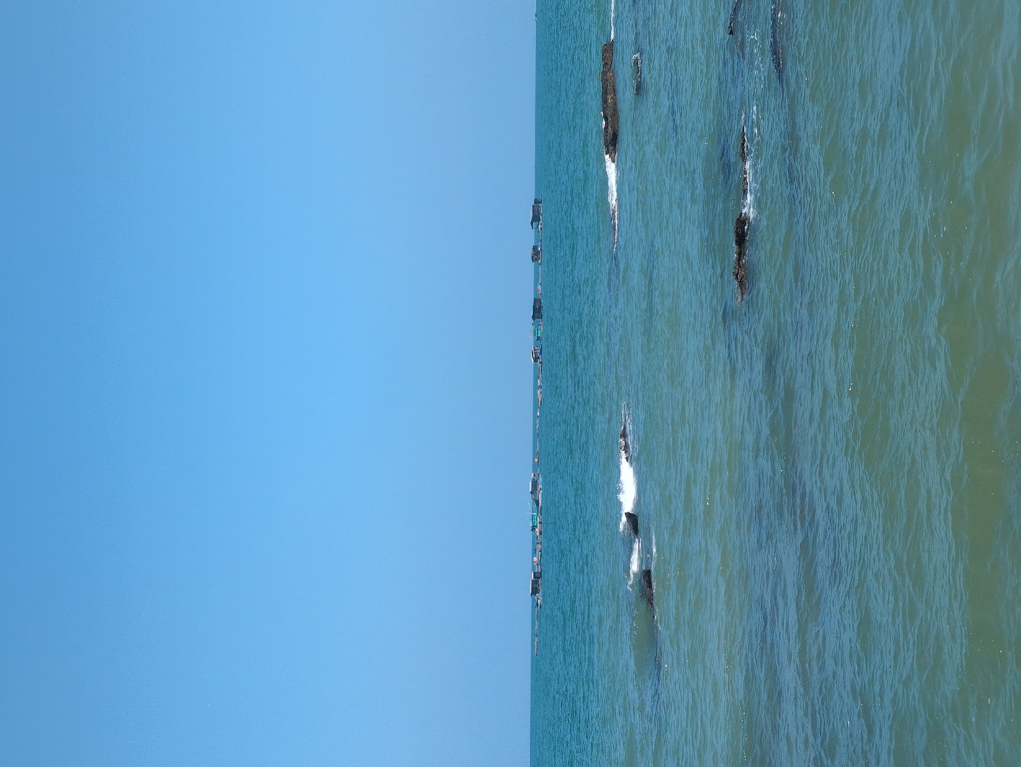 The image shows an aerial view of the sea and sky in Phú Quốc, Vietnam. The left side of the image is predominantly clear blue sky. The right side is water, with visible wave patterns and dark, presumably rocky, outcroppings dotting the surface. A vertical line of small boats is visible along the horizon separating the sea from the sky.