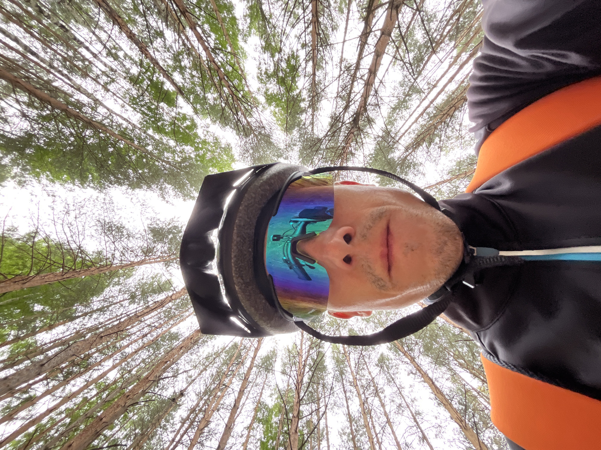 This is a low-angle, close-up, first-person perspective photograph taken outdoors in a forest environment, likely in or near the city of Chaikovsky, Russia. The image is dominated by tall, slender trees with a canopy of green leaves against a bright, overcast sky. The photographer, wearing a black helmet with reflective, multicolored goggles, is prominently featured. The goggles reflect a scene that appears to be a bicycle and part of a forest. The photographer is also wearing a black jacket with an orange stripe across the chest. The perspective suggests the photo was taken from a very low position, looking upwards through the trees, possibly while riding a bicycle. The overall impression is one of an outdoor activity in a natural setting.