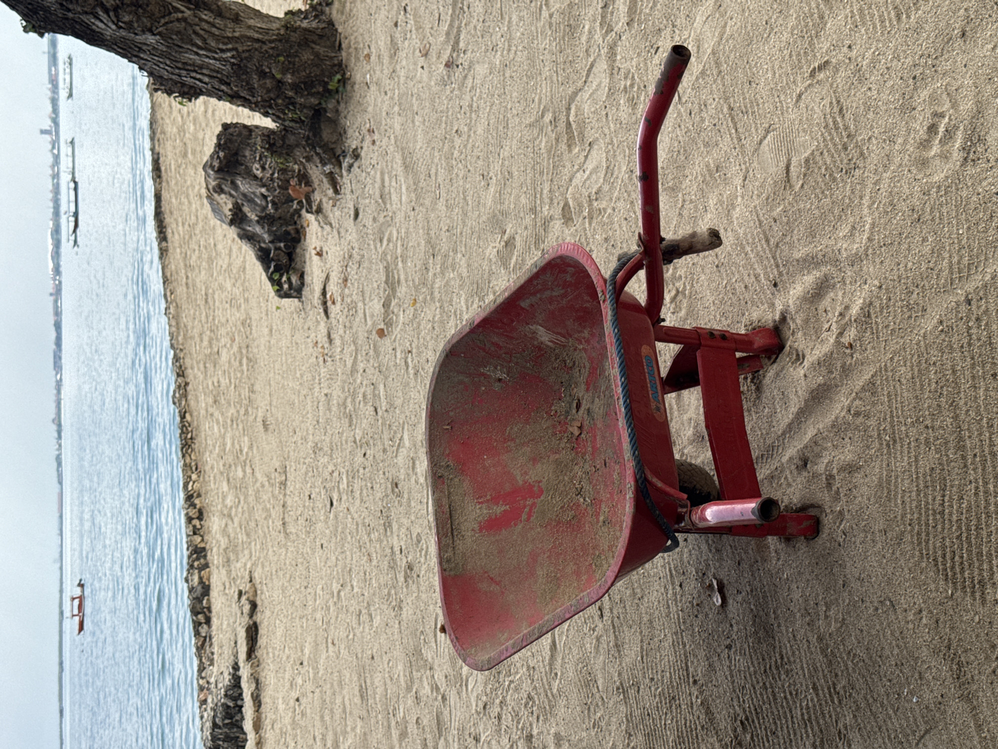 A red wheelbarrow filled with sand sits on a sandy beach in Kuta Selatan, Indonesia. The ocean is visible in the background, with small boats on the water. A tree trunk is partially visible on the left side of the image.