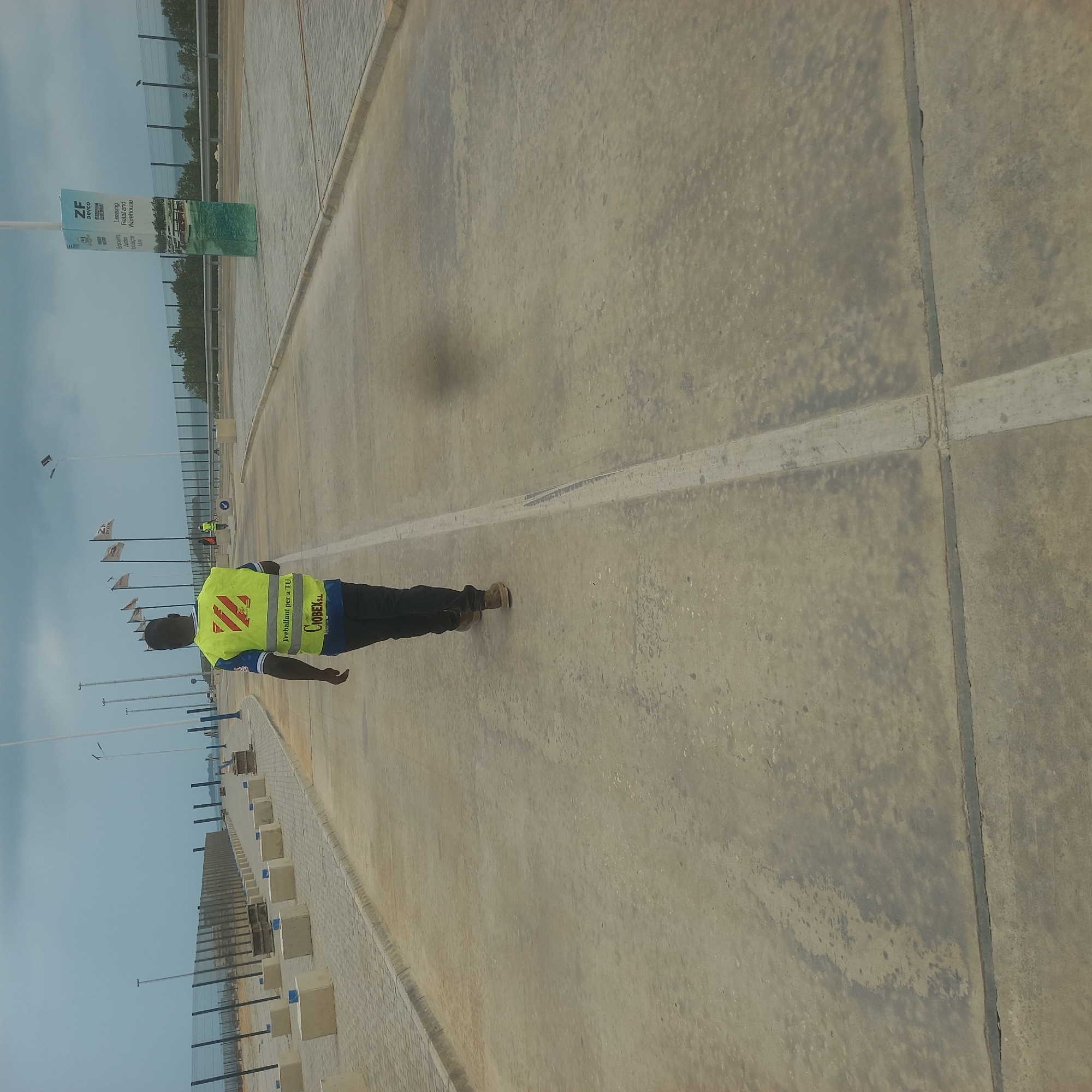 An African man wearing a bright yellow safety vest with red stripes and dark pants walks away from the camera on a concrete path. The vest has text that reads "Treballant per a TU" and "CIOBEX.SL". He is walking towards a large, modern-looking facility with a chain-link fence and numerous tall poles with lights. In the background, several flags are visible, and a large informational sign stands near a body of water. The sky is overcast, and the overall environment appears to be an industrial or port area. The location is specified as Bububu, Tanzania.