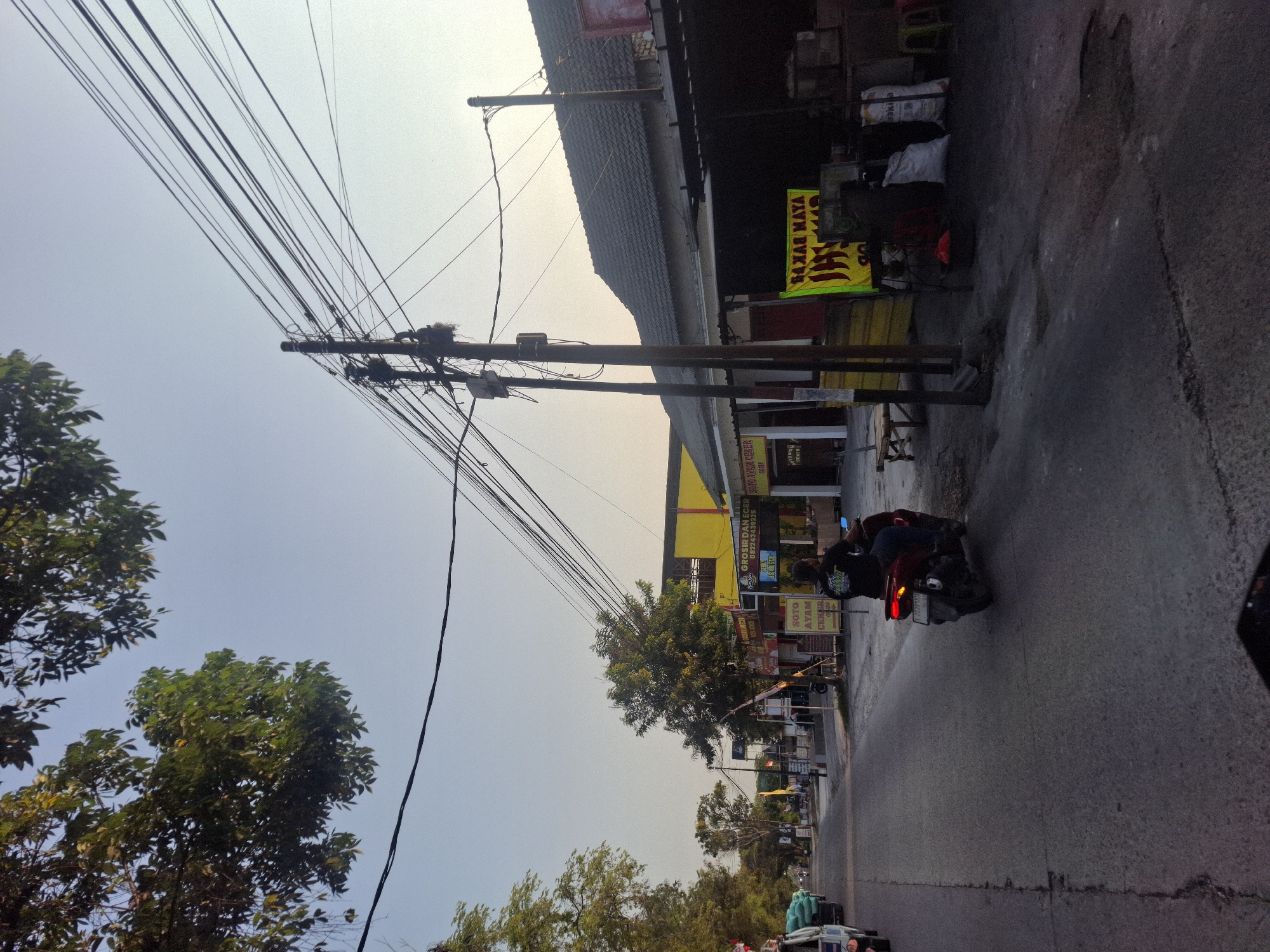 The image depicts a street scene at dusk in Sumberwudi, Indonesia. A man on a red motorcycle is riding down the asphalt road, which has some cracks and patches. The road is lined with trees on the left and buildings on the right. Overhead, a dense network of power lines crisscrosses the sky, supported by utility poles. One building on the right appears to be a shop with merchandise displayed outside, including sacks and crates. A yellow banner with Indonesian text hangs above the shop. Another building further down the street is painted bright yellow and has a prominent sign that is partially obscured. The sky is clear with a gradient of light, suggesting the sun is setting or has recently set. The overall impression is of a typical Indonesian street with active commerce and essential infrastructure.