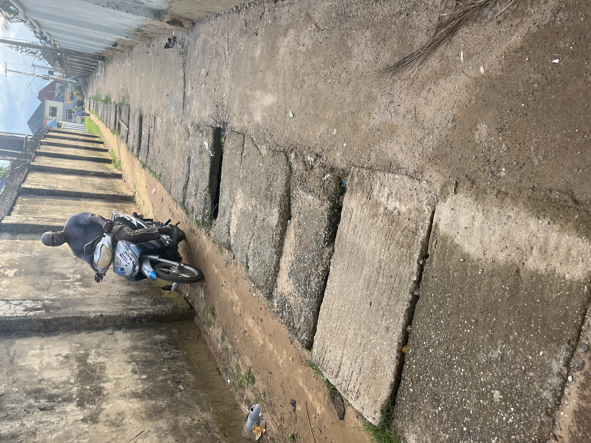 The image depicts a narrow, unpaved alleyway or access path in an urban environment, consistent with areas found in Abuja, Nigeria. The path is bordered by tall concrete walls on both sides, which appear to be property fences.
In the foreground and mid-ground, a dark-skinned adult male is seated on a grey and blue scooter, facing forward. He wears a grey short-sleeved shirt and dark trousers. His hands are on the handlebars, and his feet are near the ground, suggesting he is either stationary or moving slowly. The scooter features a front headlight and a visible front wheel.
The ground surface consists of multiple elements. To the left of the scooter is a dirt path alongside the left concrete wall, with sparse green vegetation and some visible litter, including a discarded plastic bottle. The central portion of the path is covered by a series of uneven, rectangular concrete slabs, many of which are cracked, broken, or misaligned, revealing gaps and an underlying dirt or debris-filled trench. To the right of the scooter and extending into the mid-ground, the path is primarily dirt and gravel, showing patches of moisture and scattered debris, including a dried broom made of sticks.
Both boundary walls are constructed of concrete. The left wall exhibits significant weathering and discoloration. The right wall appears somewhat lighter in color, and sections near the top are fitted with barbed or razor wire.
In the background, the alley extends into the distance, narrowing and curving slightly. At the far end, several multi-story buildings with pitched roofs are visible against an overcast sky. A lone individual is discernible walking away from the camera in the distance. Utility poles with overhead wires are also present in the background, further indicating an urban or suburban setting.