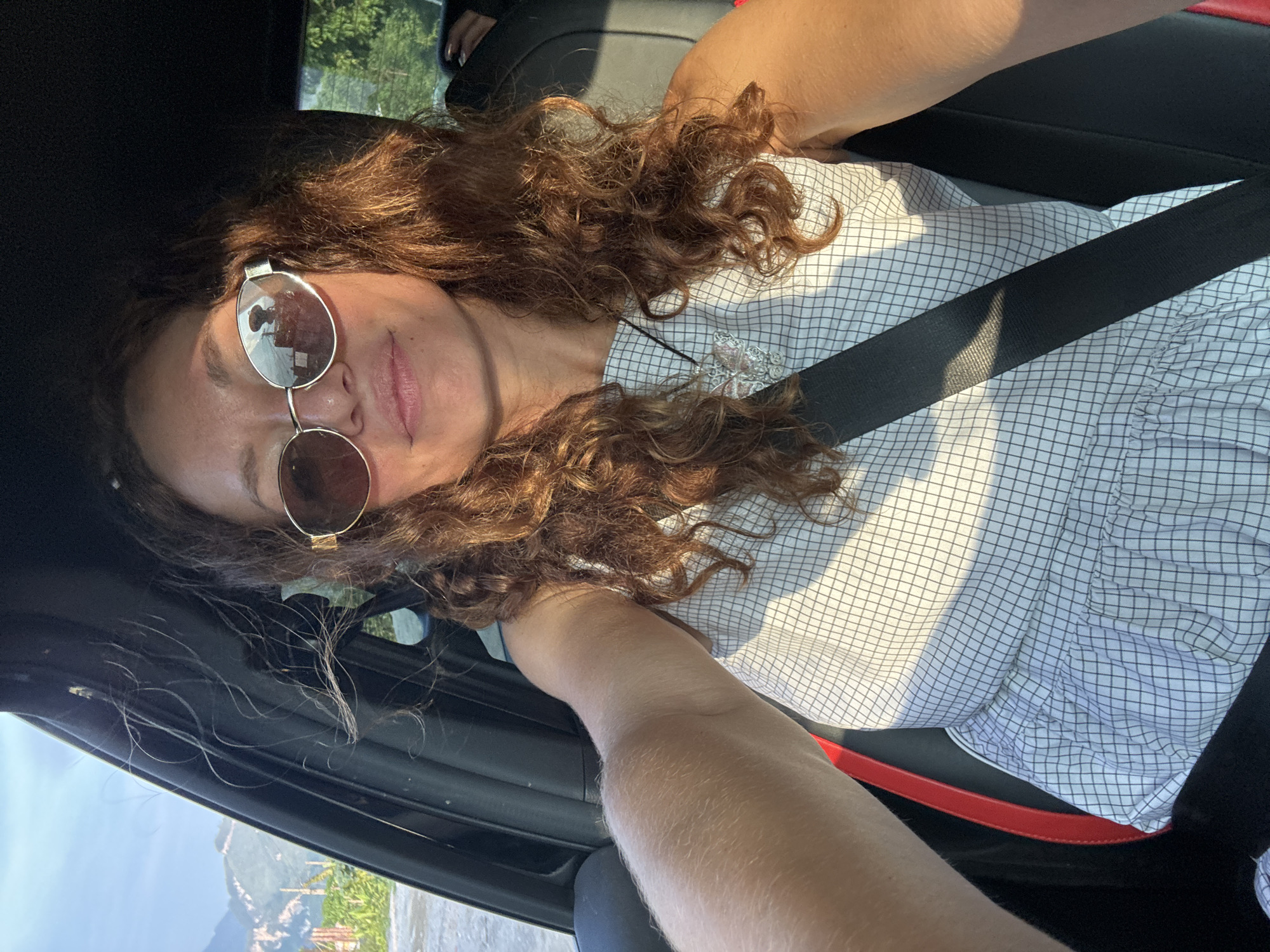 The image is a selfie taken inside a vehicle, likely a car, showing a person wearing sunglasses and a seatbelt. The person has curly brown hair and is wearing a light-colored, checkered shirt. The vehicle's interior is visible, including parts of the dashboard and door. Outside the car window, a glimpse of a landscape is visible, featuring what appears to be trees and mountains. The specific location is Du Già Commune, Vietnam.