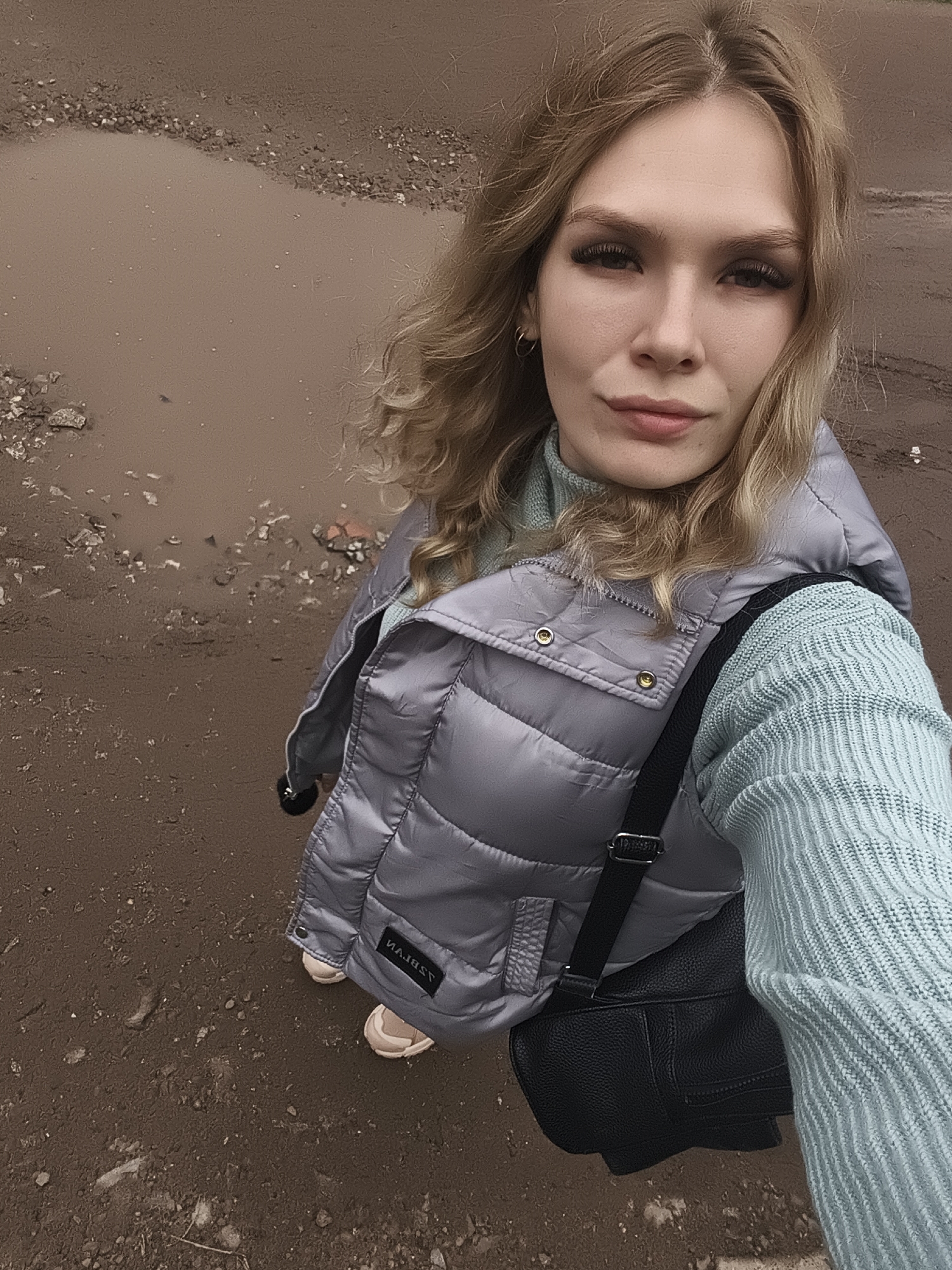 A young woman with blonde, wavy hair is captured in a selfie, looking directly at the camera with a neutral expression. She has prominent eyelashes and appears to be wearing light makeup. She is dressed in a light blue, ribbed turtleneck sweater and a silver-grey puffer vest, across which a black textured shoulder bag is slung. Light-colored sneakers are visible on her feet.
The setting is an outdoor, unpaved area characterized by muddy, brown ground and several murky puddles. Small stones and debris are scattered across the terrain. The diffused lighting suggests it is daytime, likely under overcast skies, with the wet ground indicating recent rain or damp conditions.
A black patch with mirrored white text, possibly "MAJEST", is visible on her puffer vest. The scene depicts a casual moment in a somewhat rugged, possibly undeveloped or neglected environment in Izhevsk, Russia.