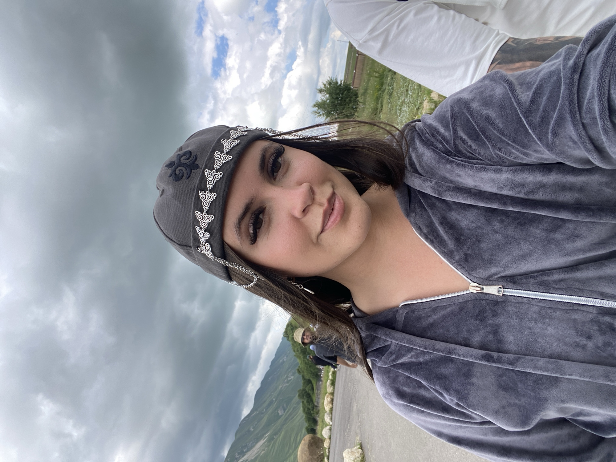 A young woman with dark hair and heavy eye makeup poses for a selfie in Tsalkita, Russia. She is wearing a gray velour hoodie and a dark gray beanie adorned with silver chain detailing and an embroidered emblem. The woman is smiling slightly and looking towards the camera. In the background, a vast, green mountainous landscape stretches under a cloudy sky. Two other individuals are visible in the middle ground, one wearing a hat and a dark shirt, and another person to the right of the main subject is partially visible, wearing a white shirt. The ground in the foreground appears to be a paved area with scattered rocks.
