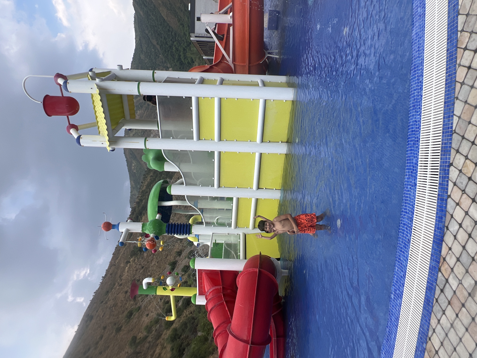 In Khumsan, Uzbekistan, a young boy stands waist-deep in a bright blue swimming pool in front of a multi-colored water park structure. The water park features red slides, yellow panels, and white support columns, with a red bucket at the top of a central tower. In the background, arid, brown hills rise steeply under a cloudy sky. The boy is wearing orange patterned swim trunks and is facing the camera with his arms raised. Another child is partially visible behind him in the water. The pool's edge is lined with blue and white tiles, and the surrounding area is paved with textured stone tiles.