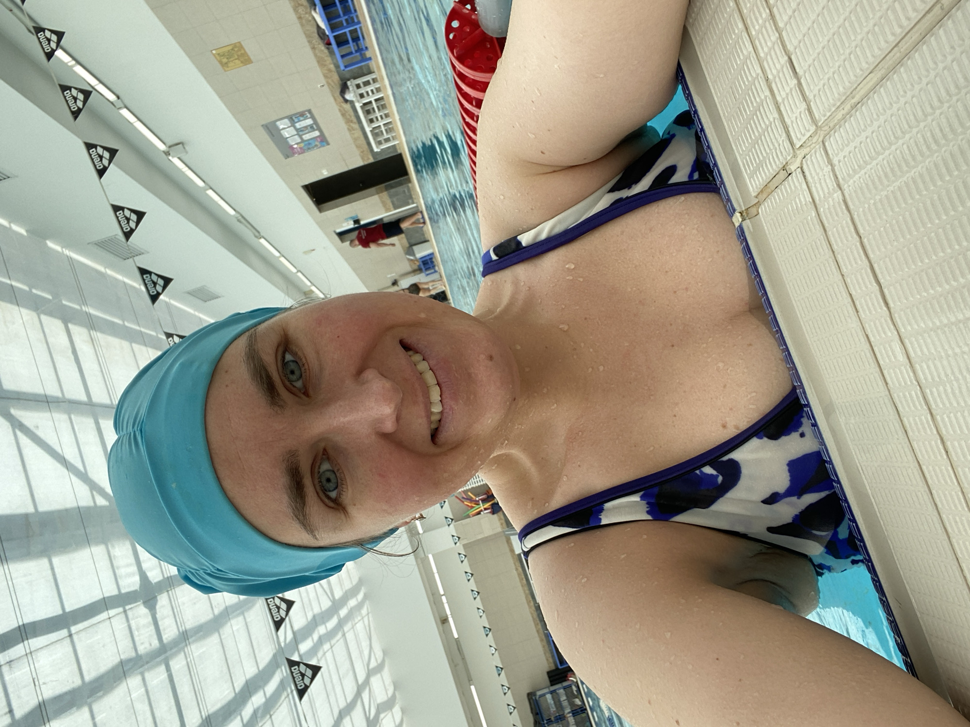 A woman is taking a selfie at the Yekaterinburg Swimming pool. She is wearing a blue swimming cap and a black and white swimsuit. Water droplets cover her skin. She is at the edge of the pool with white tiles. In the background, there is a pool with blue water, lane markers, and other swimmers. Arena branded flags line the ceiling.