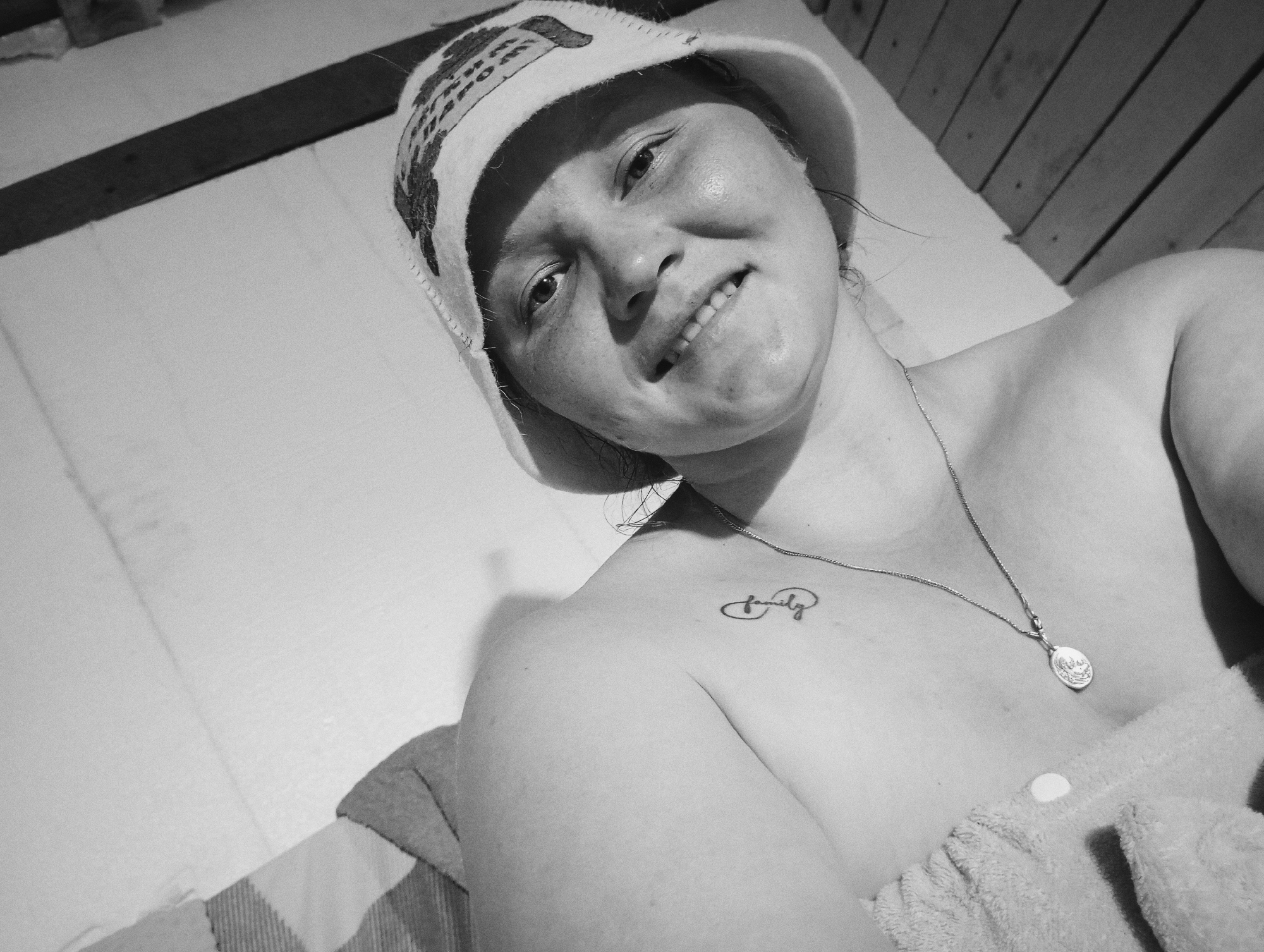 A grayscale, high-angle selfie shows a woman wearing a patterned hat and a towel around her chest. She is lying down with her head tilted back and a slight smile on her face. A delicate silver necklace with a pendant hangs around her neck. There is a small tattoo of the word "family" in an infinity symbol on her left shoulder blade. The background consists of wooden planks and a white surface. The image is indoors, possibly in a sauna or spa, in the city of Ryabki, Russia.