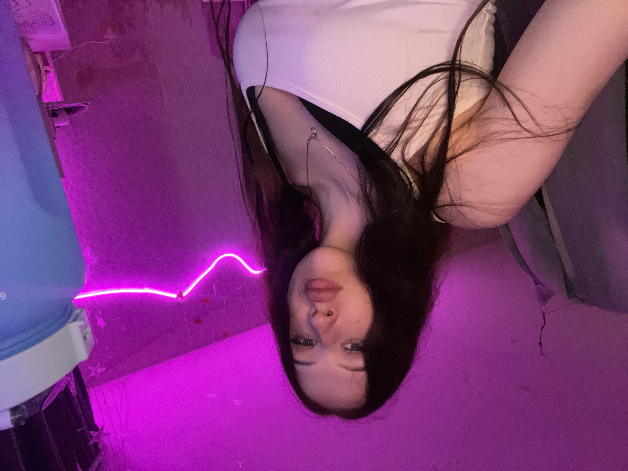 The image is a selfie taken indoors, likely in a bedroom, with predominantly pink lighting. The subject is a young woman with long, dark hair, looking directly at the camera. She is wearing a white tank top over a black bra and a silver necklace. To the right of the frame, a large blue water bottle is visible. The background features a wall with some decorative elements, including a neon pink light strip shaped like a wavy line, and small red heart decals. The overall impression is of a casual, personal space. The location is not specified within the image.