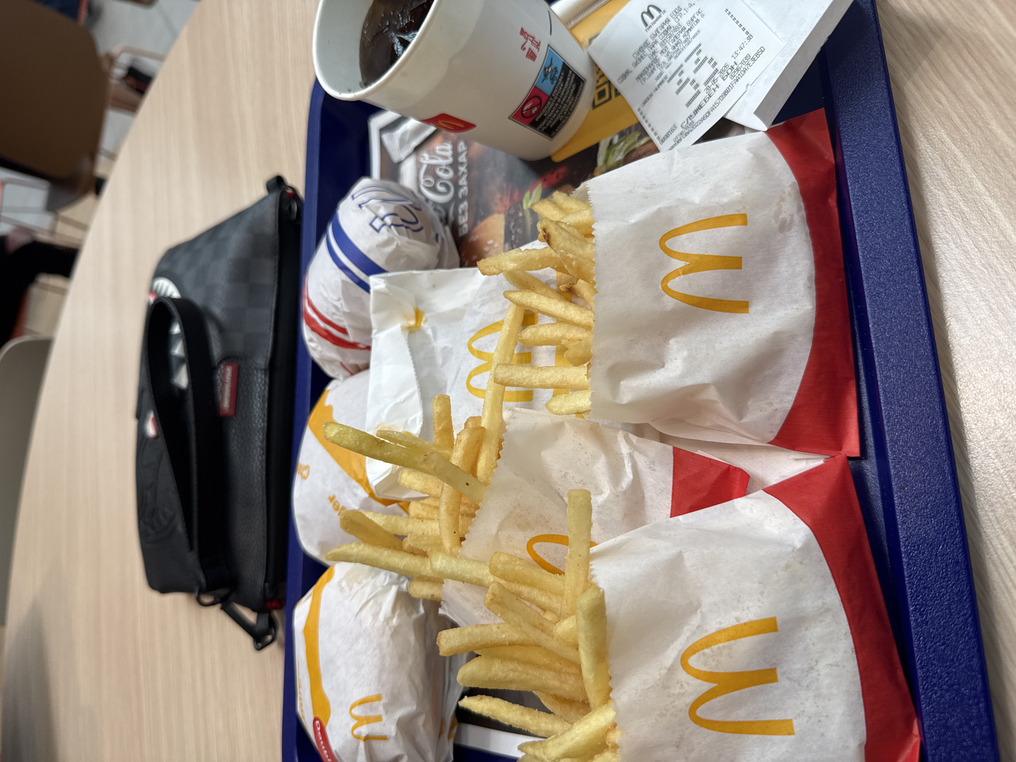 A tray of McDonald's food sits on a light brown table. The tray, lined in red, contains a cup of Coca-Cola Zero Sugar, a hamburger in a wrapper, and three packets of french fries. The receipt, issued by "McDonald's Mall Gallery Burgas, blvd. Yanko Komitov B" in Burgas, Bulgaria, lays on top of the food. A black and grey checkered backpack sits next to the tray on the left side of the frame. The restaurant is located in Sofia Business Park.