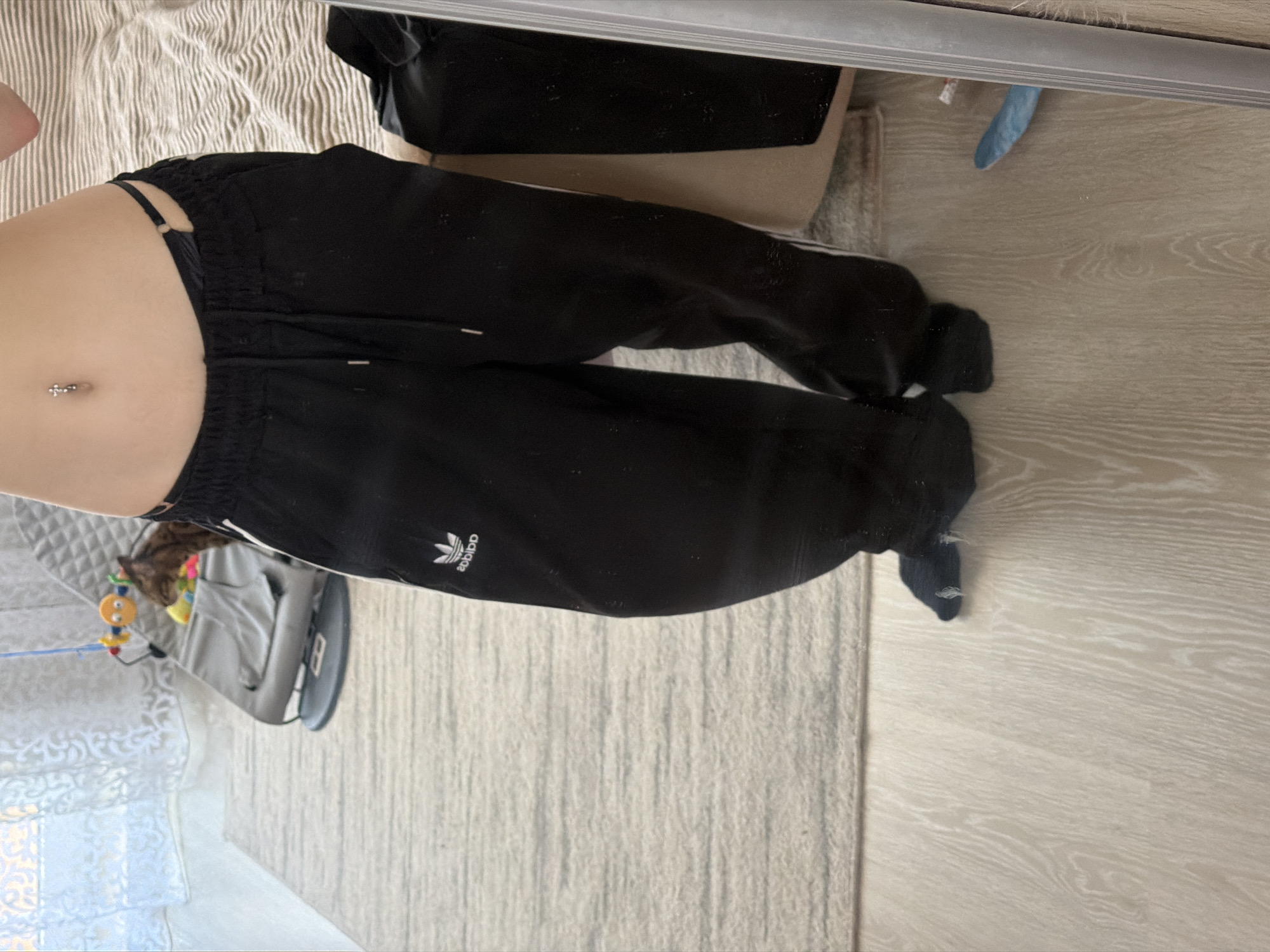 The image shows a torso wearing black Adidas sweatpants with a navel piercing. The person is reflected in a mirror, with a bed and a baby bouncer with toys visible in the background. The floor is light-colored laminate.
There is no evidence to determine the location to be Noviy, Russia.