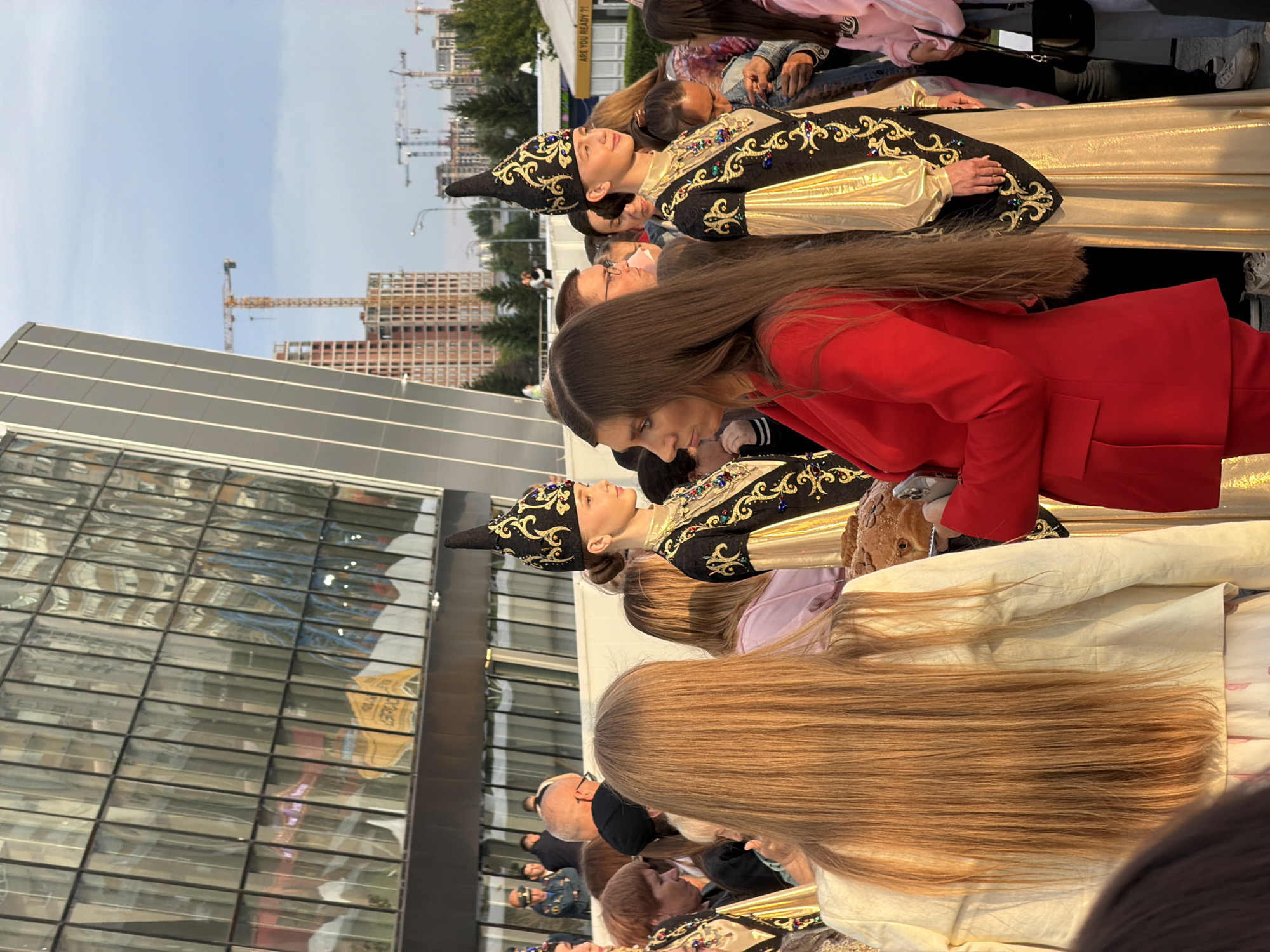 In an outdoor setting, a group of people are gathered, with several individuals dressed in elaborate traditional costumes. One person wears a black and gold outfit with intricate embroidery and a tall, pointed hat. Another person is seen in a bright red blazer, and others are in casual clothing. The background features a modern building with a glass facade, reflecting the surroundings. Beyond the building, there are construction cranes and a sky with some clouds, suggesting a partially overcast day. The people appear to be participating in or observing an event. The lighting suggests it could be late afternoon or early evening. One visible sign in the reflection of the glass building reads "ARE YOU READY".