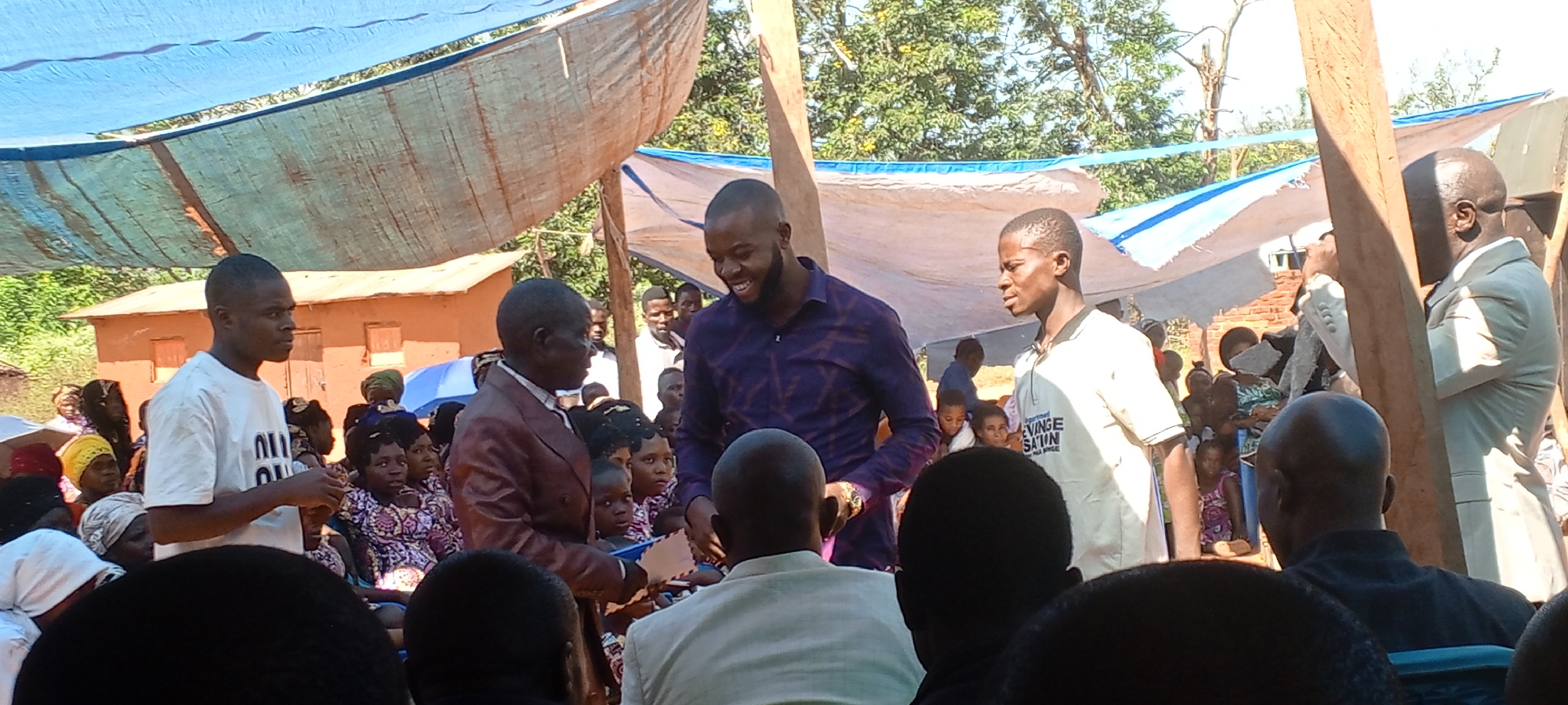 The image depicts an outdoor event in a rural setting, likely in or near Kalemie, Democratic Republic of the Congo. A large group of people, including men, women, and children, are gathered under a makeshift canopy constructed of blue and white tarpaulins. Several individuals stand in the foreground, suggesting they are the focus of the event. One man wears a maroon suit and appears to be holding a document or item presented to him by another man in a purple patterned shirt who is smiling. To their right stands a man in a light-colored shirt with writing on it. Other adults and children are seated in the background, some wearing patterned dresses and head coverings. The environment shows a combination of structures and natural elements, including a brick building, trees, and vegetation. The lighting is bright, indicating the event is occurring during the day.