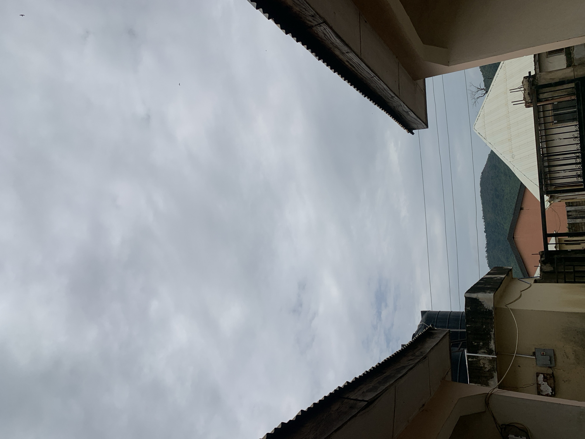 A low-angle shot taken from between buildings in Lokoja, Nigeria. The sky is overcast with gray clouds. The edges of two buildings with corrugated metal roofs frame the top and bottom of the image. Power lines are visible stretching across the open space. In the distance, another building with a white, slanted roof is seen, with a small hill or mountain visible behind it. A water tank is visible on the roof of the building at the bottom of the frame. The building exteriors appear weathered.