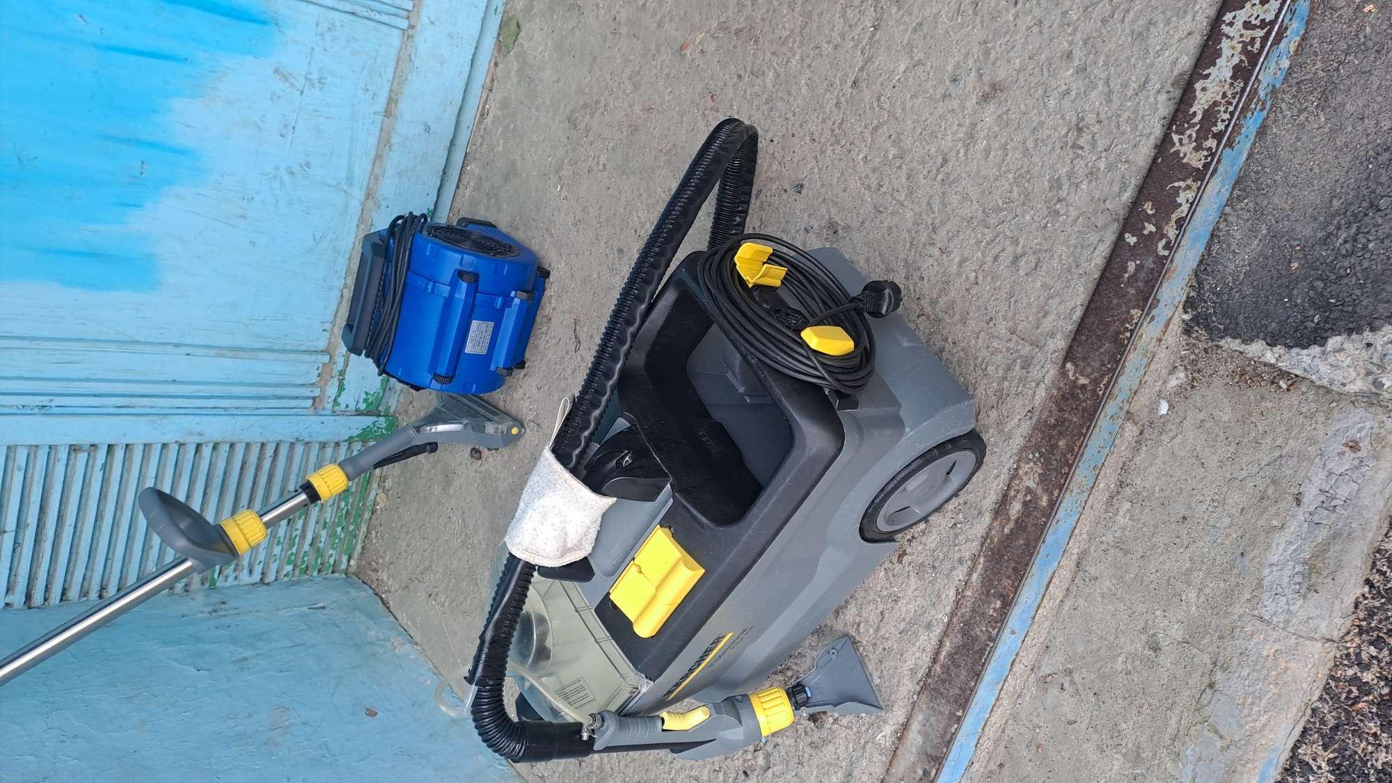 An outdoor, eye-level, full shot shows a grey and black Karcher carpet cleaner with a grey hose and yellow accents, as well as a blue blower fan, resting on a concrete surface in front of a blue, weathered wooden door. A silver and grey cleaning wand with yellow accents is propped up to the left of the carpet cleaner, its nozzle resting on the concrete. The Karcher carpet cleaner is a rectangular unit with wheels on the bottom, a grey body, a black top with a clear tank and yellow storage compartments, and a black hose wrapped around the front. A white cloth is draped over the top of the carpet cleaner. The blue blower fan is a rectangular device with a black power cord coiled on top and a black grille on the front. The blue wooden door has horizontal planks and is peeling in several places. The concrete surface shows wear and tear with cracks and patches of darker material.