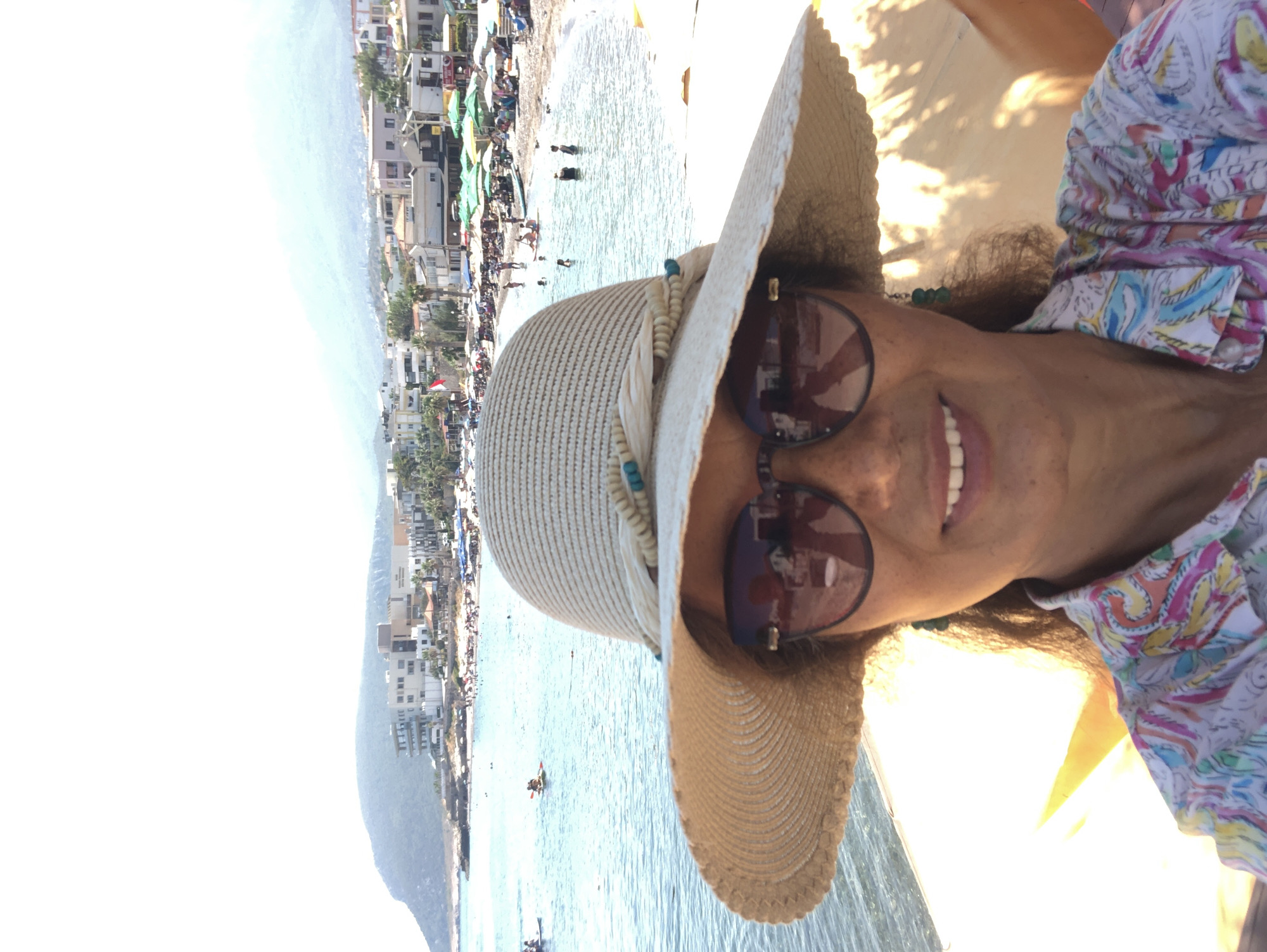 In this close-up selfie, a woman with dark curly hair, wearing a wide-brimmed straw hat adorned with turquoise beads, sunglasses with dark lenses, and a colorful paisley-print shirt, smiles at the camera. Her reflection is visible in her sunglasses, showing a hint of the camera she is holding. She is on a beach, with the turquoise sea and a hilly coastline dotted with buildings behind her. Several people are visible in the water, some swimming, others on paddleboards. Beach umbrellas dot the sandy shoreline in front of the buildings. The scene appears to be located in the city of Karaburun, Turkey, a coastal town known for its beaches and scenic landscape. The lighting suggests a sunny day.